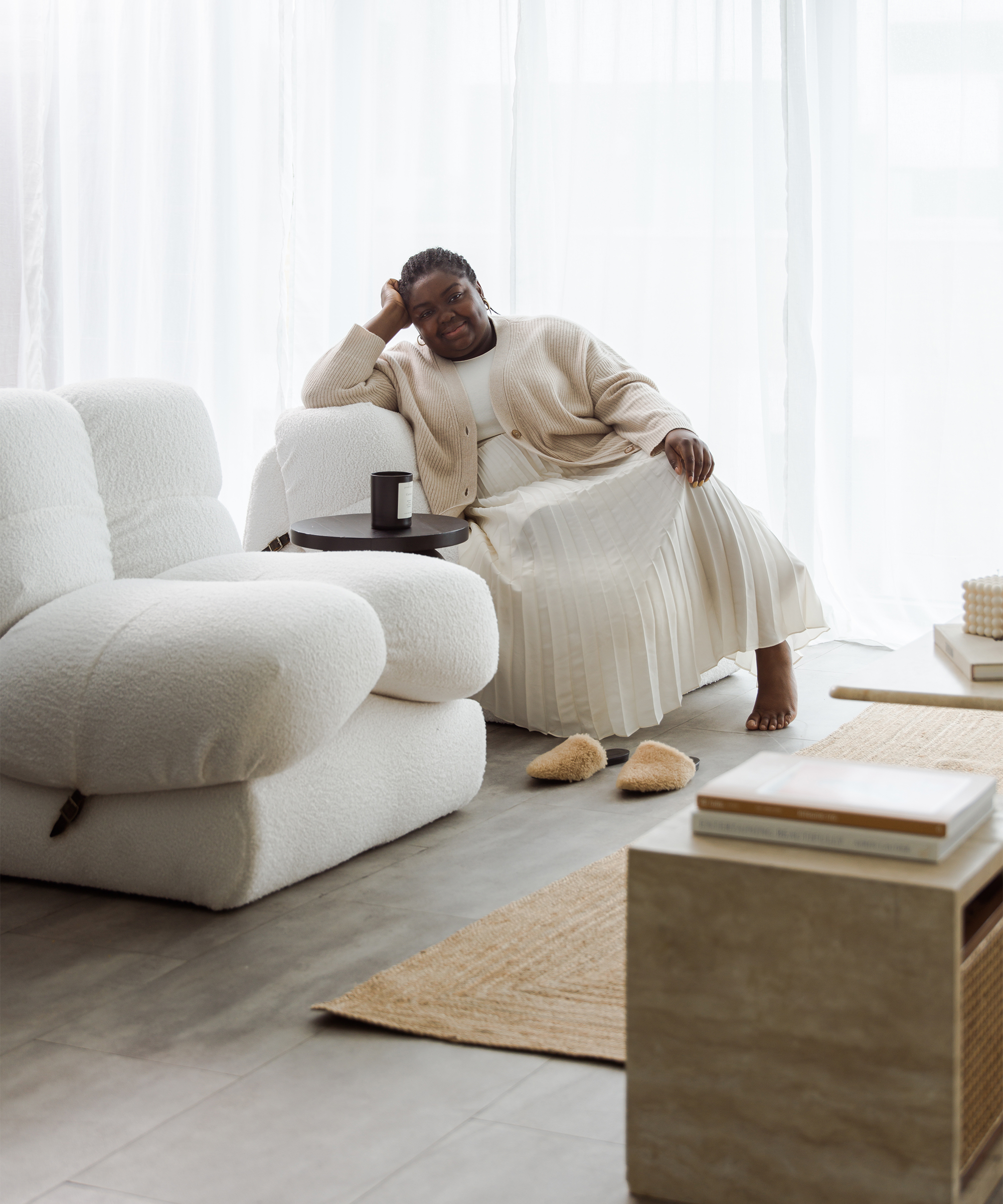 A woman in a cream outfit sits relaxed on a white armchair in a bright, modern living room styled by Abisola Omole. She smiles, resting her head on her hand, with slippers nearby and sunlight streaming through sheer curtains.