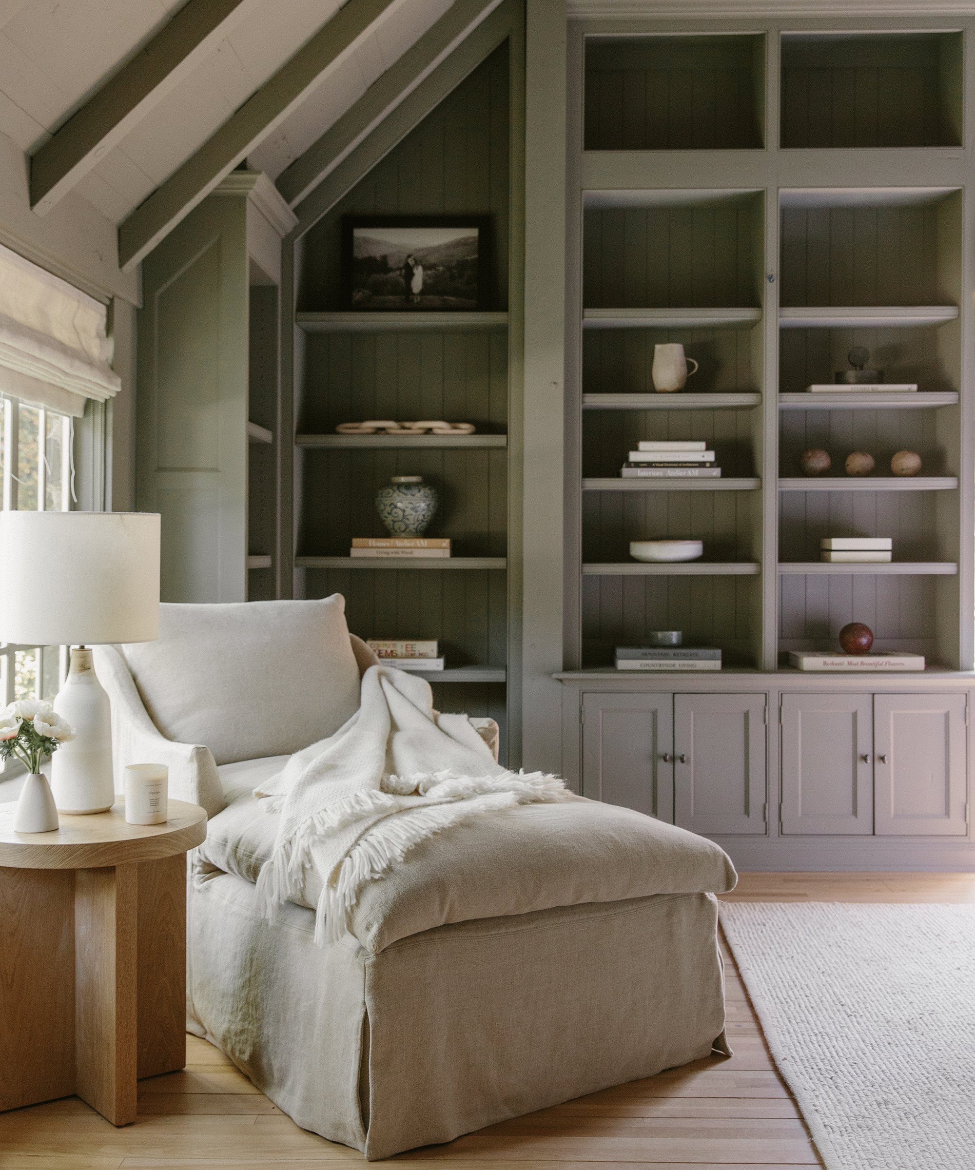 A cozy reading nook with a cushioned armchair and ottoman, draped with a fringed throw, sits beside a round wooden side table. Built-in shelves and books create a serene backdrop, echoing the relaxed elegance of Jenni Kayne interiors.