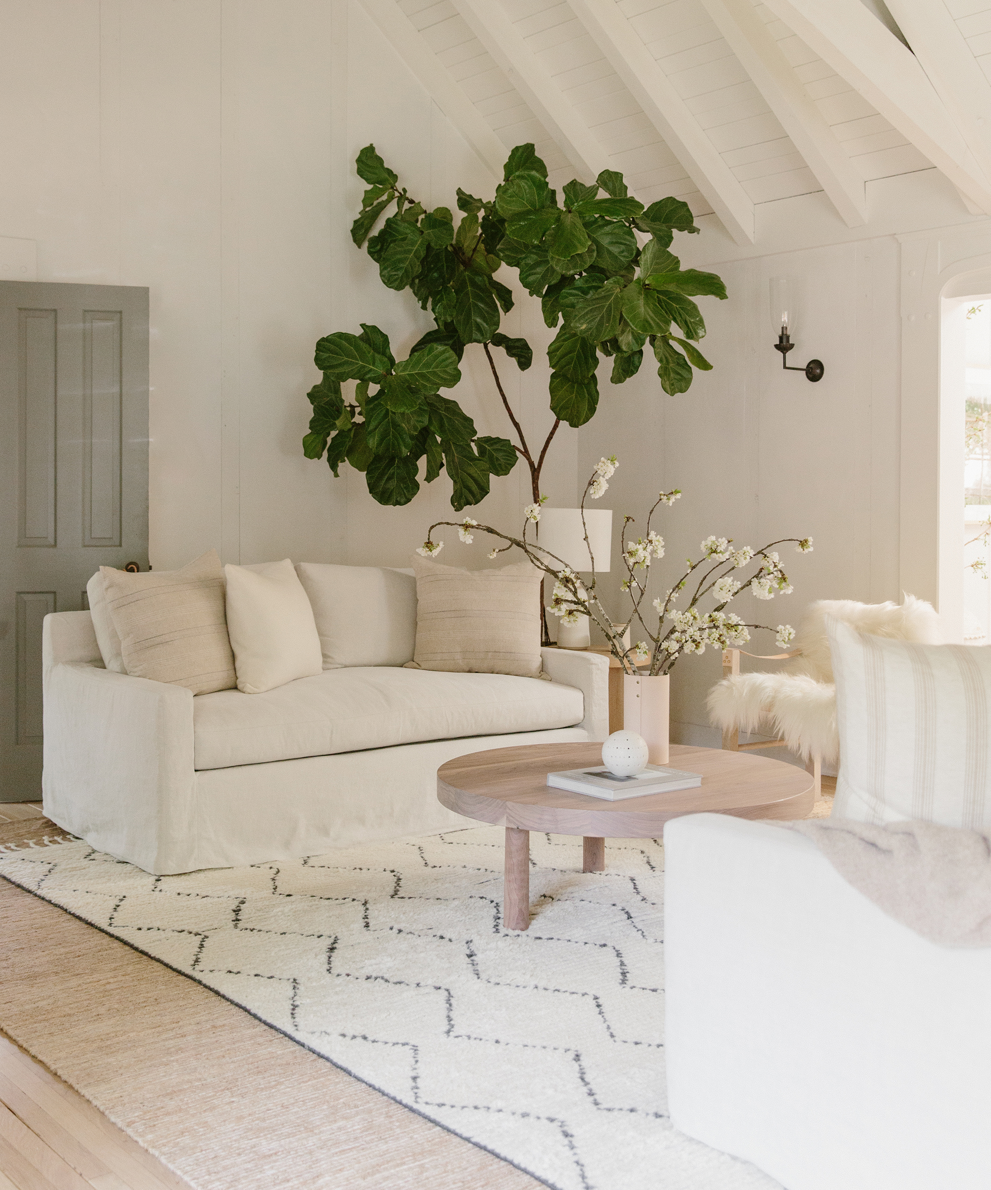 Bright, airy living room remodel with a neutral sofa, light wood coffee table, patterned rug, large potted plant, and a vase with flowering branches. White walls and high vaulted ceiling create an open, serene atmosphere.