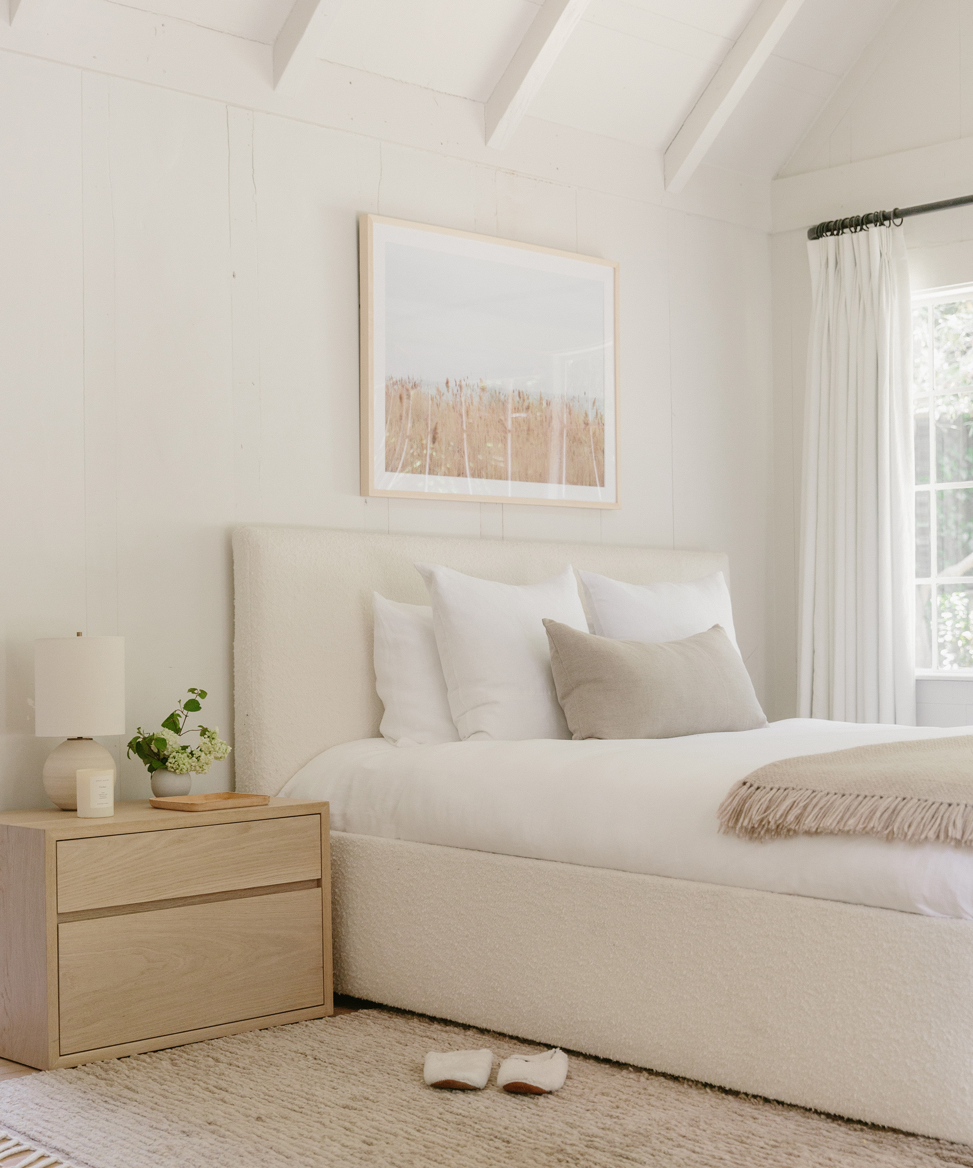 A cozy, minimalist bedroom remodel features a white bed, neutral pillows, light wood nightstand, lamp, plant, beige throw, slippers on the rug, and nature-themed artwork on the white wall. Sunlight pours in from a window.