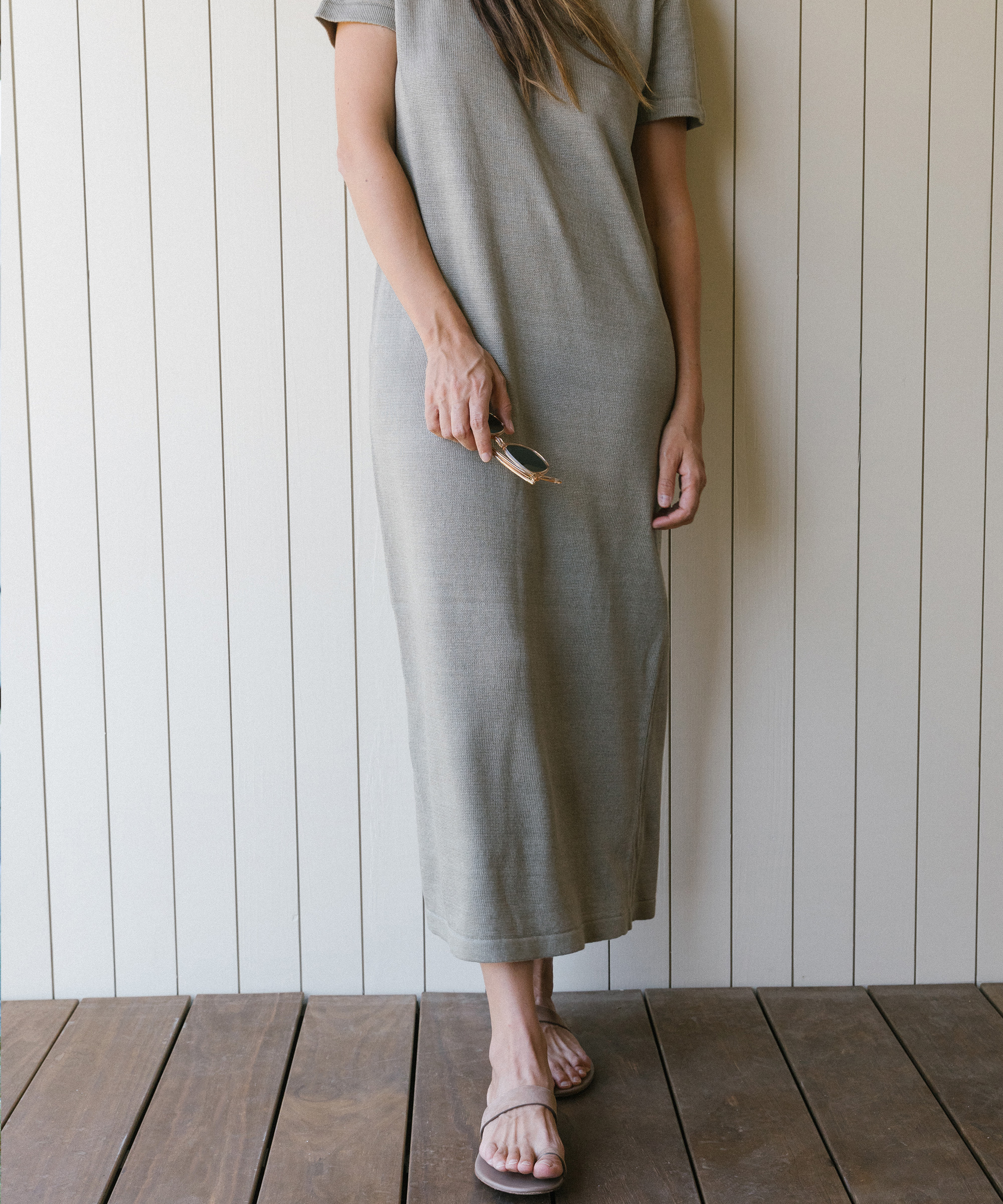 A woman in a gray, short-sleeve, ankle-length dress stands on a wooden floor, holding sunglasses in one hand—an effortless choice for Labor Day outfits. She wears light-colored sandals, and the background is a white paneled wall. Her face is not visible.