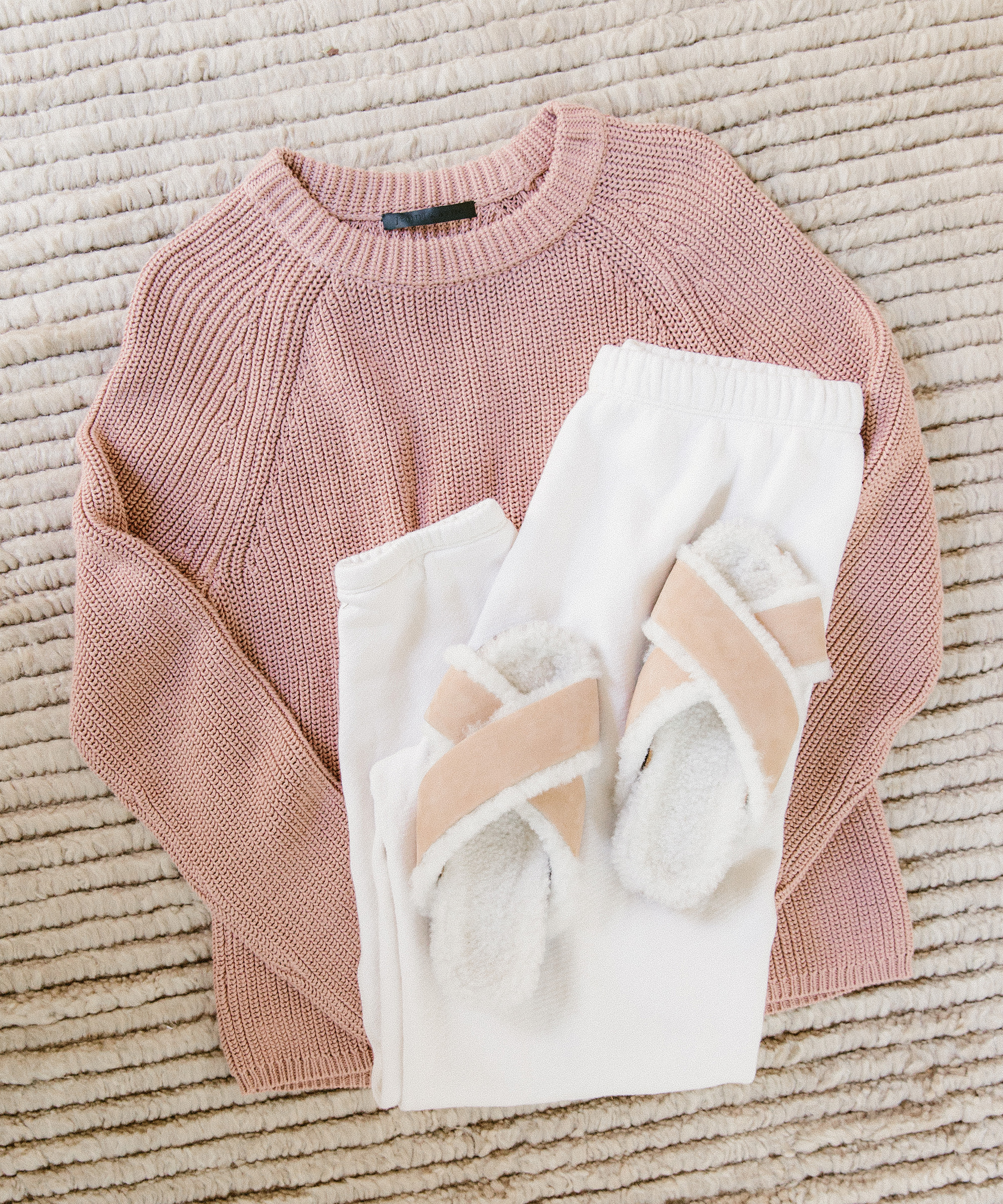 A folded pink knit sweater, white sweatpants, and beige fuzzy slippers are neatly arranged on a textured cream rug—perfect staples to mix and match in your summer capsule wardrobe.