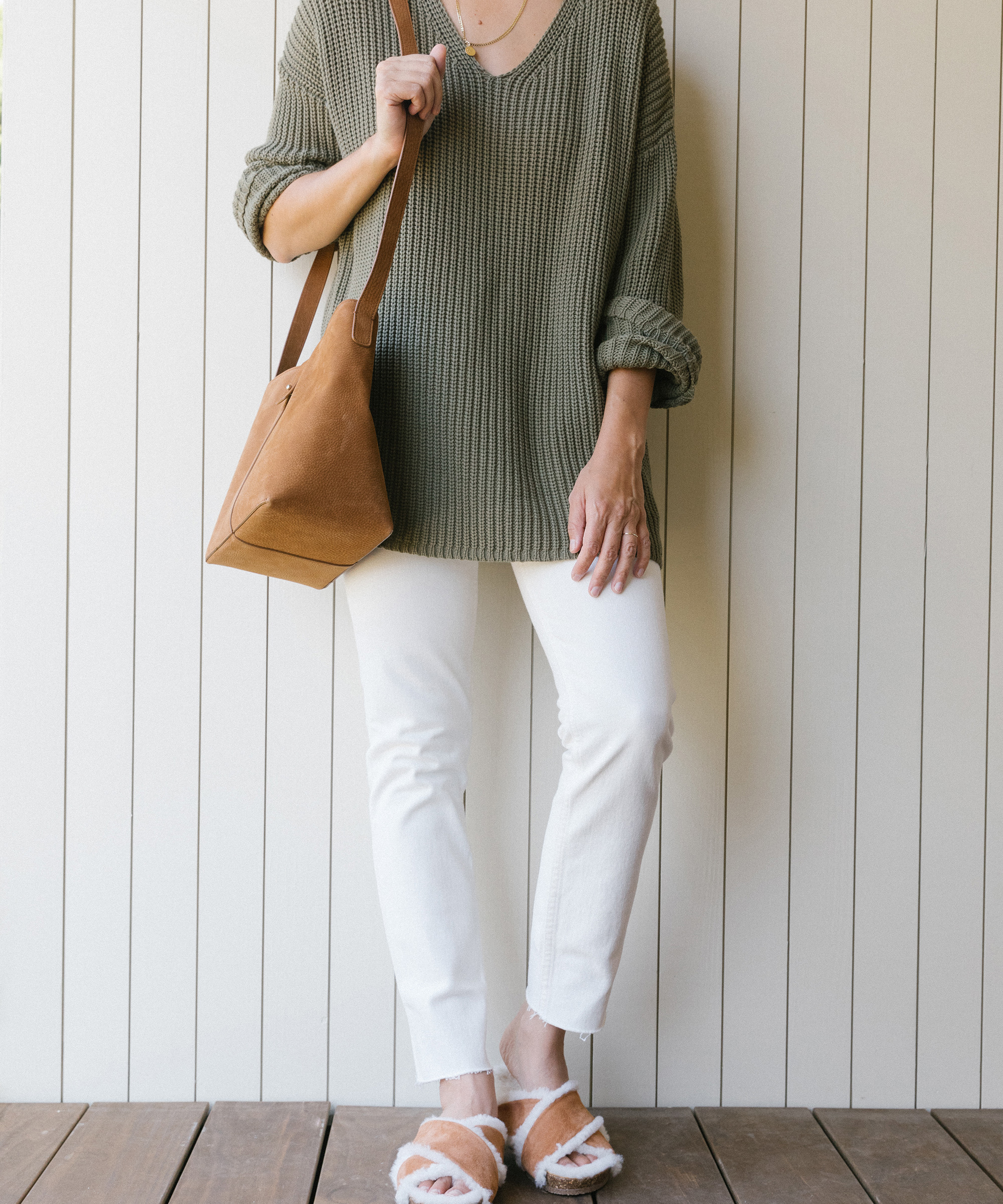 Kate Brien standing against a white wall wearing white jeans, cotton cabin sweater, and shearling crossover sandal with bag around shoulder.