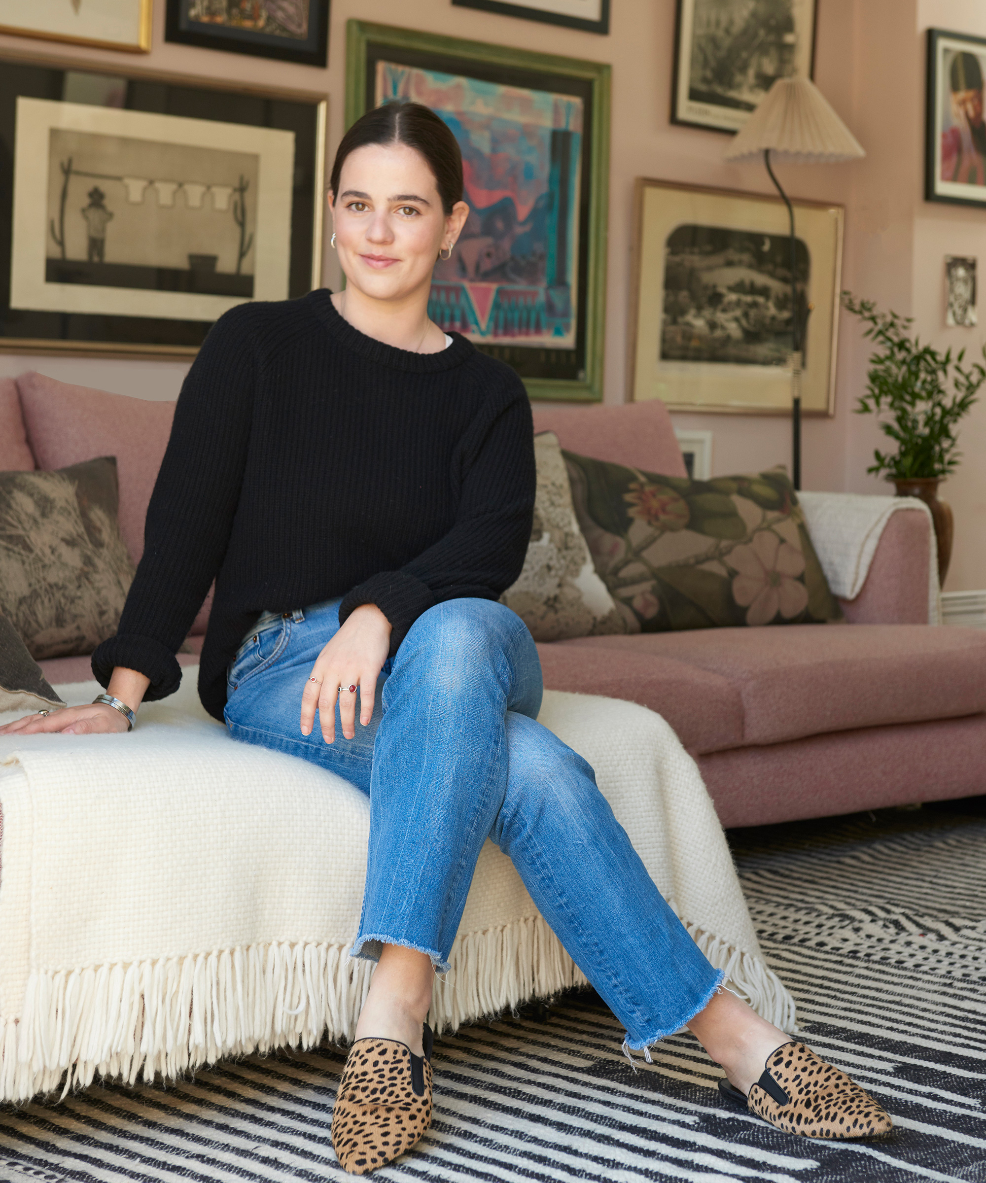 A woman with dark hair in a bun sits on a beige sofa covered with a white fringed blanket. She wears a black sweater, blue jeans, and leopard print shoes. Sascal Studio artwork and cushions decorate the cozy, stylish room.
