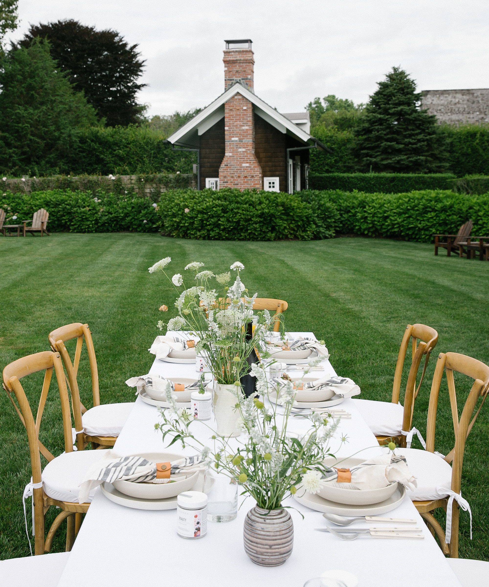 A long outdoor dining table with wooden chairs is set with white plates, napkins, and wildflower centerpieces on the lawn at Reform Club Amagansett. In the background, a small brick house nestles among lush green trees and bushes.