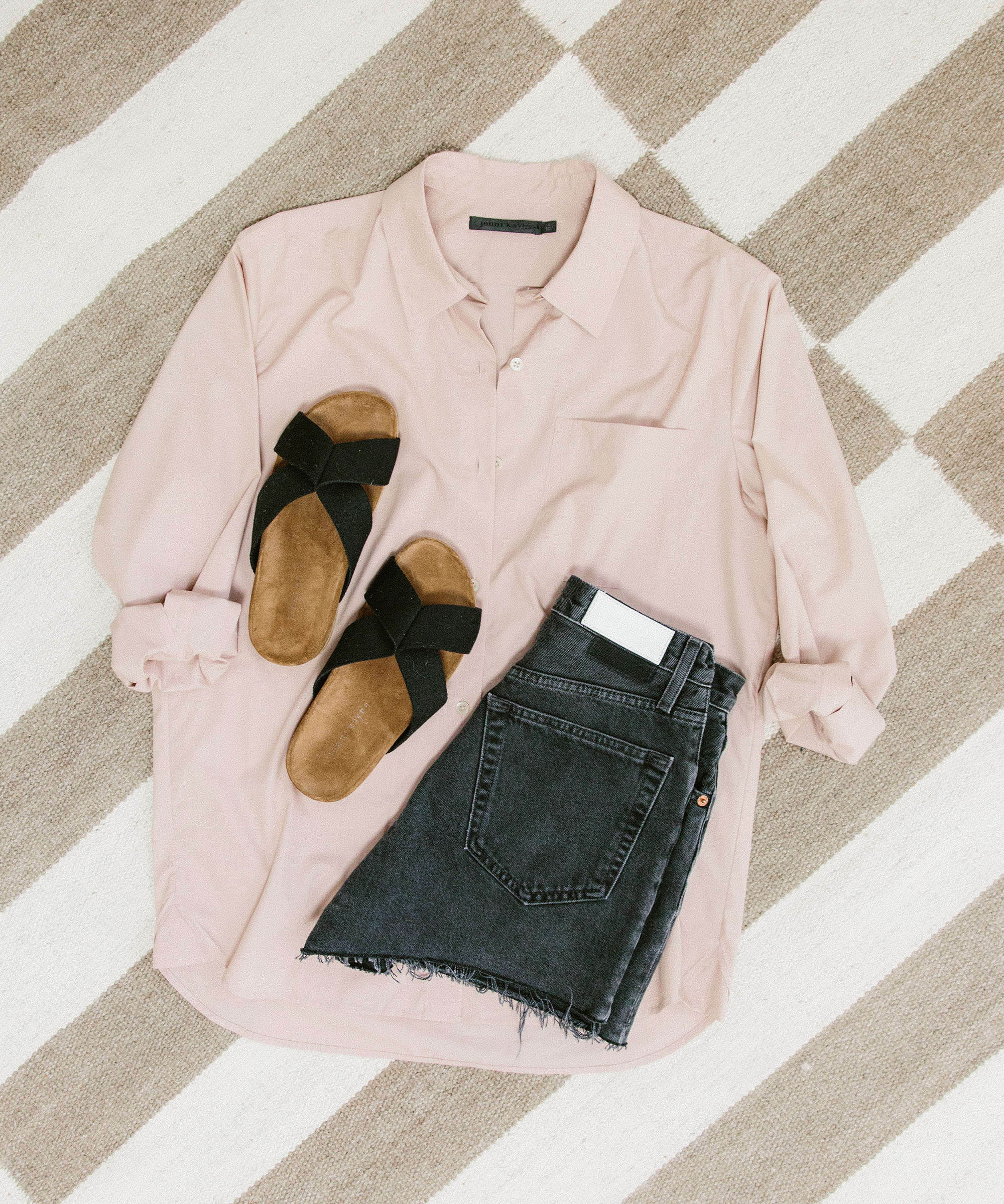 A light pink button-up shirt, black denim shorts with a frayed hem, and brown sandals with black straps are neatly arranged on a striped rug—an effortlessly chic look for your summer capsule wardrobe.