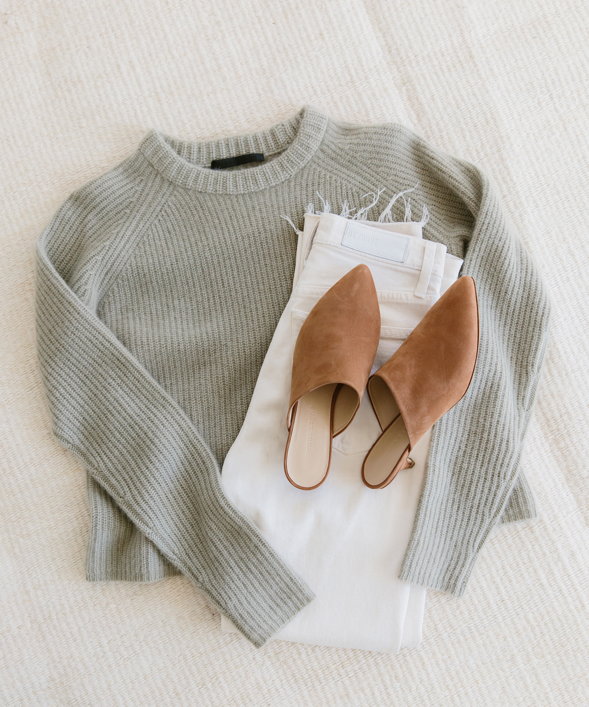 A light sage green knit sweater, white jeans, and brown pointed-toe slip-on flats are neatly arranged on a light beige textured surface—perfect pieces for any summer capsule wardrobe.