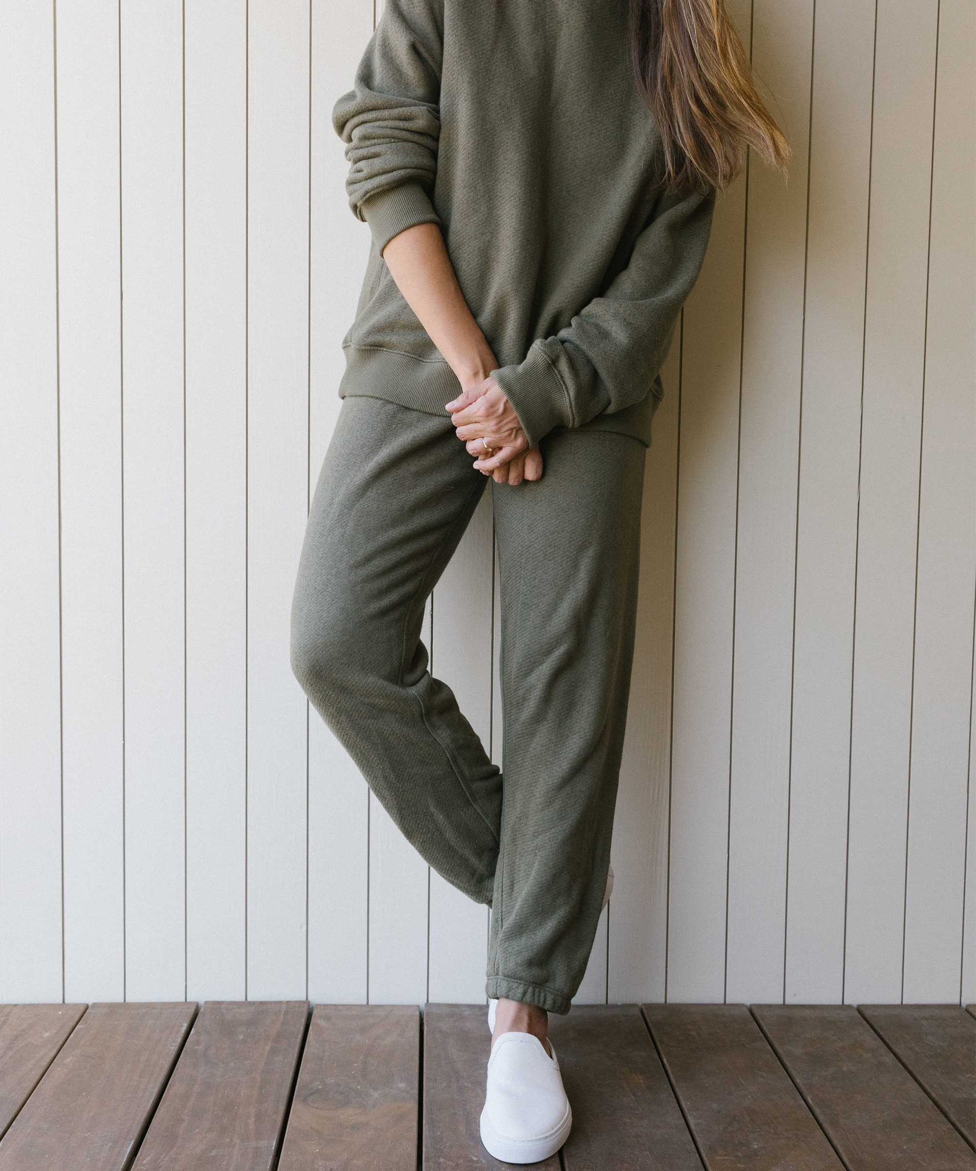 Person wearing an olive green sweatshirt and matching sweatpants stands on a wooden floor against a light-colored paneled wall; arms crossed, head not visible. These relaxed separates make for effortless Labor Day outfits, finished with white slip-on shoes.