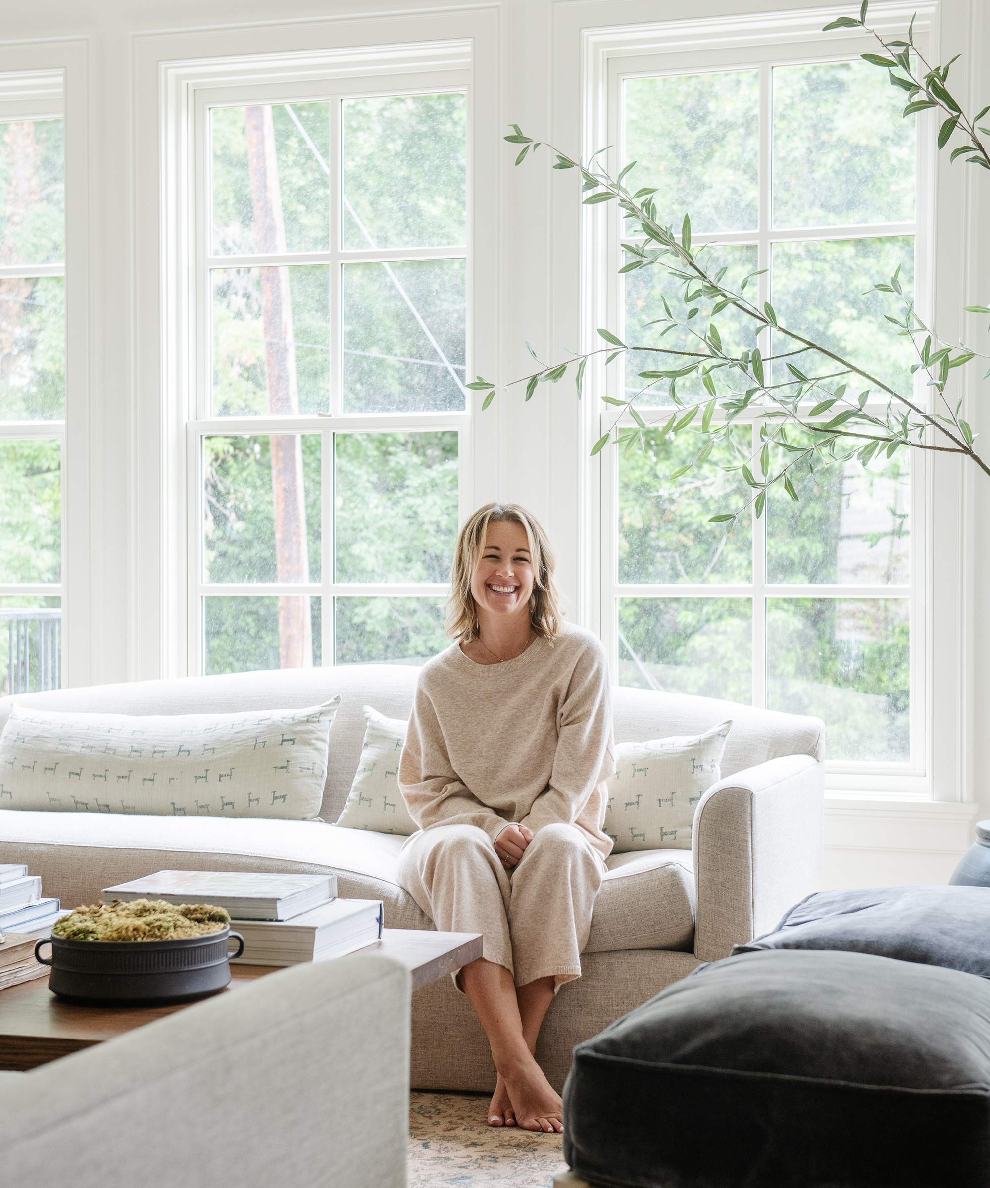 A woman in cozy loungewear sits barefoot and smiling on a light-colored sofa in a bright living room by w design collective, with large windows, leafy green views, and soft, neutral decor.