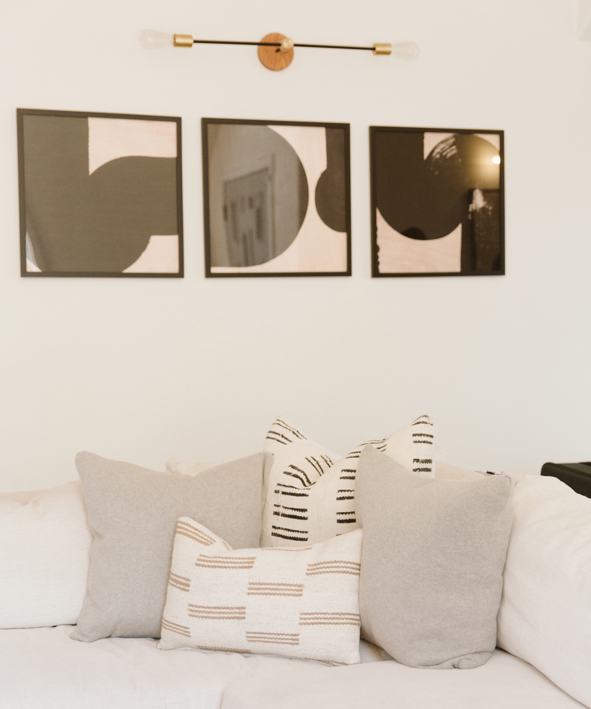 A neutral-toned living room inspired by Jaclyn Johnson features a white couch with gray and patterned pillows. Above, three abstract black-and-white framed artworks hang, while a minimalist wall sconce provides elegant light.