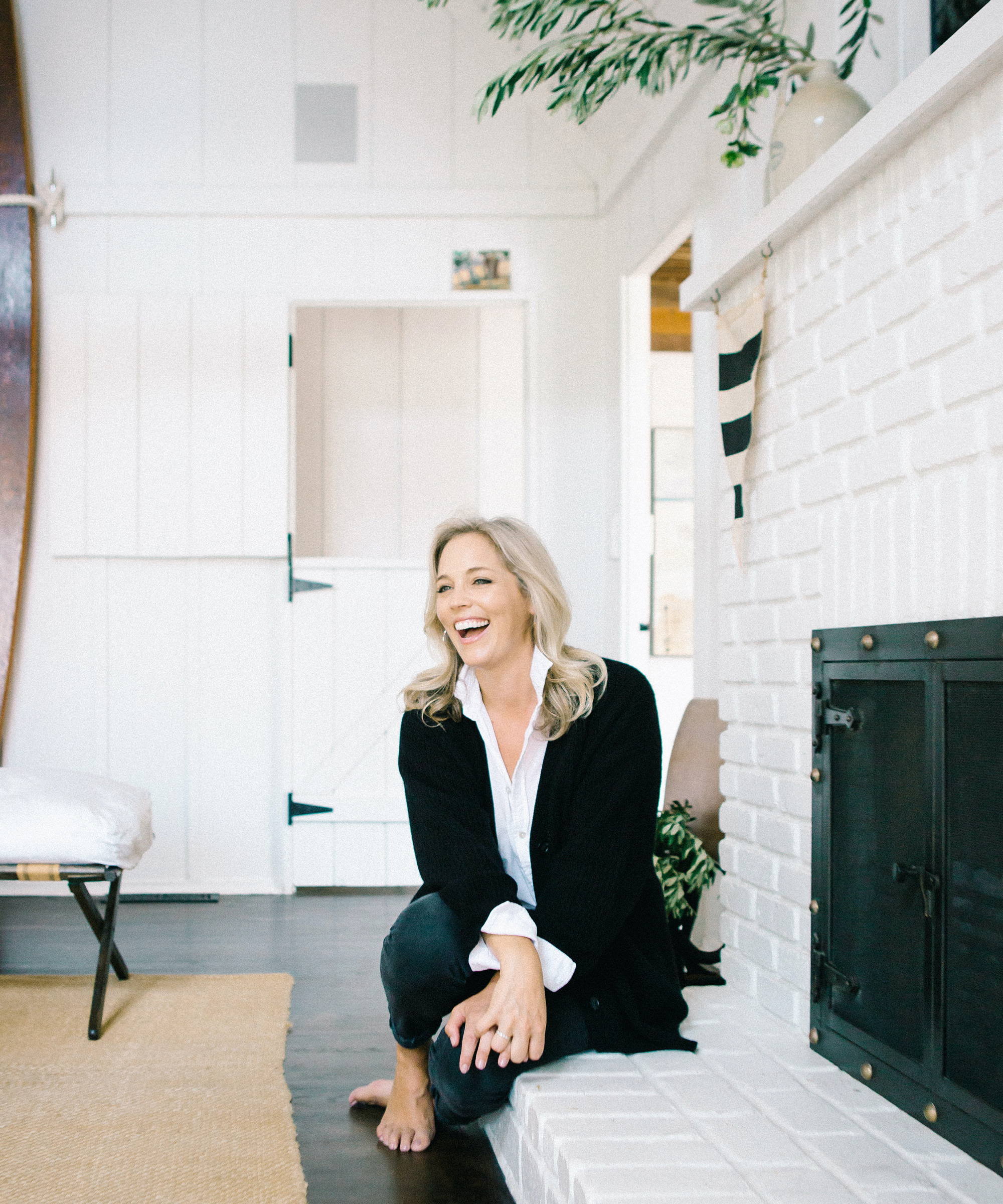 A smiling woman with blonde hair sits barefoot on the edge of a white brick fireplace in a bright, cozy living room with white walls and wooden accents, showcasing the warm, inviting style of Alexis Garrett Design.