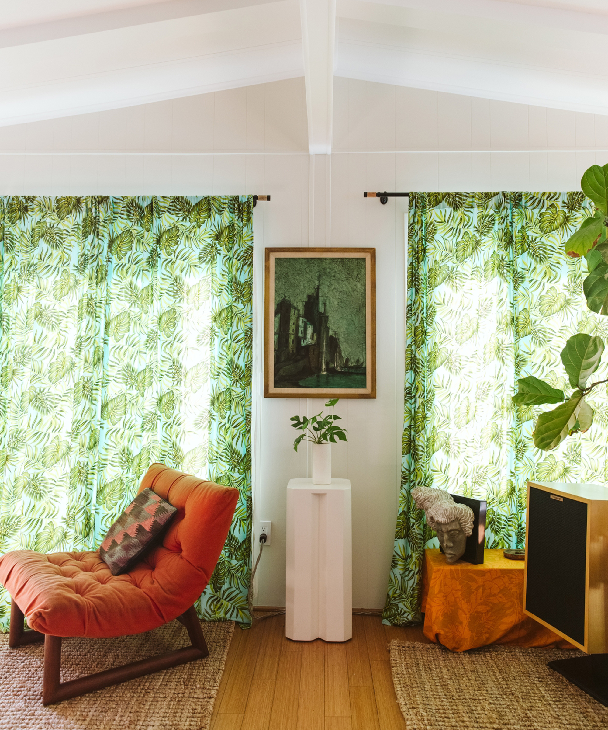 A cozy living room with a red cushioned chair, leafy green curtains, a framed painting by Marc Gabor, a tall plant, a small white pedestal with a potted plant, a speaker, and natural woven rugs on a wood floor.