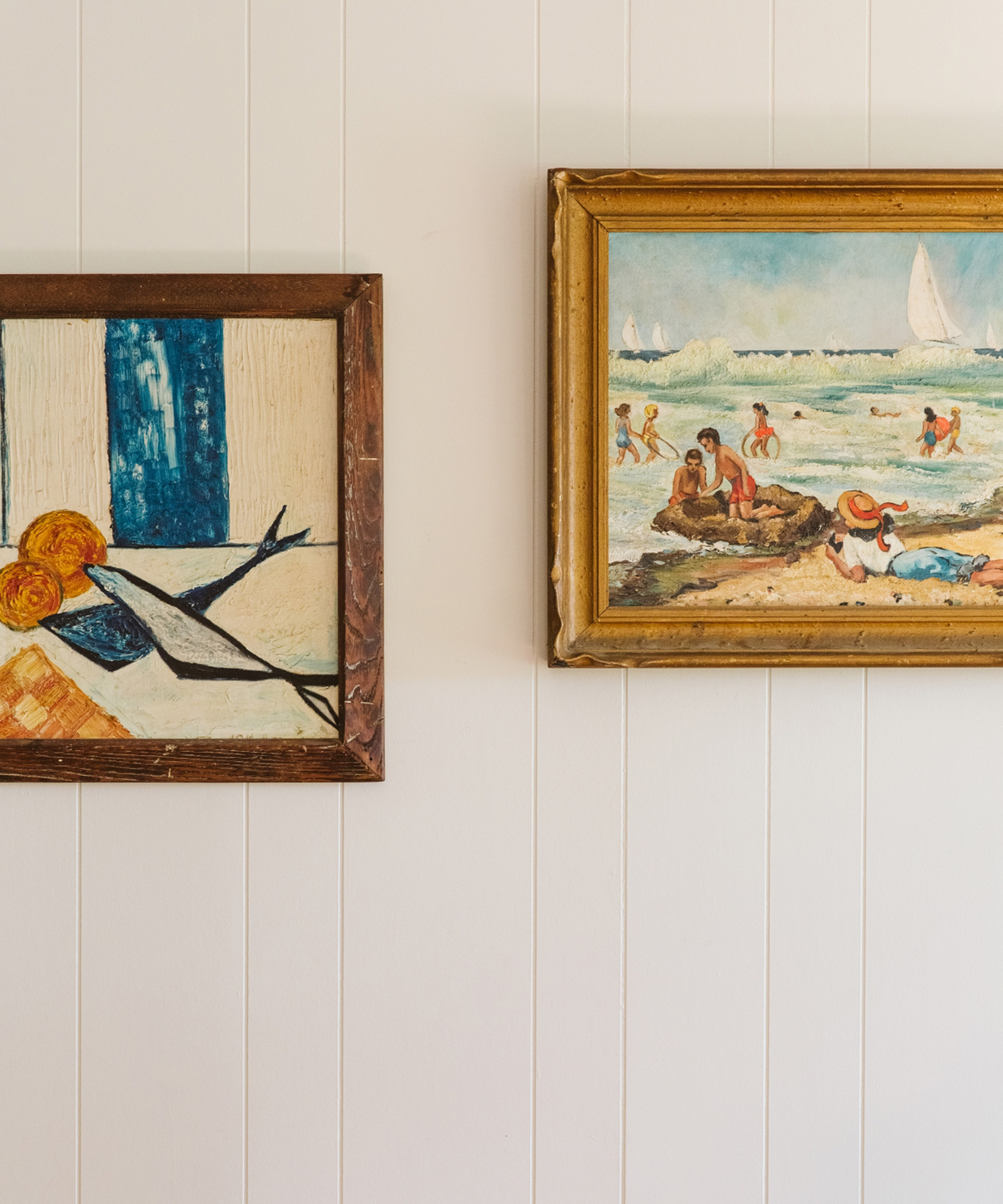 Two framed paintings hang on a white paneled wall—one an abstract piece with bold colors and shapes, the other a seaside scene with people and sailboats, evoking a Marc Gabor-inspired blend of vibrant artistry and tranquil coastal charm.
