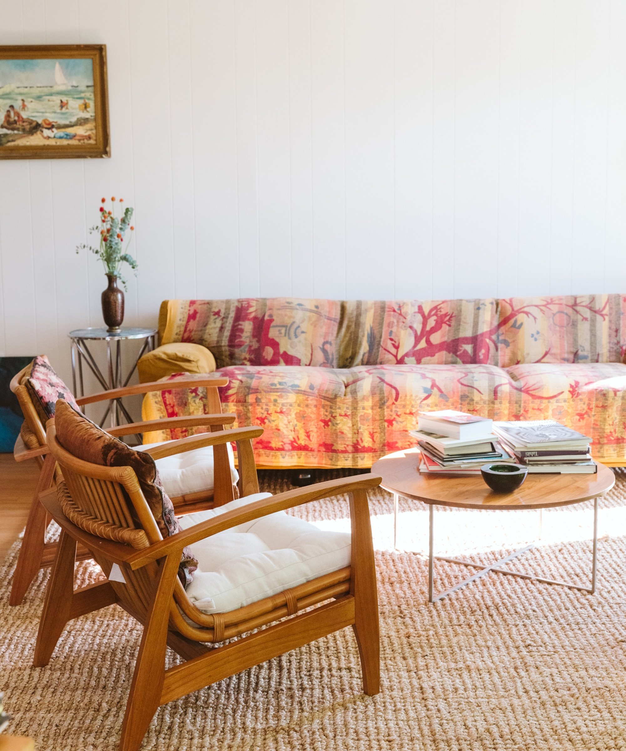 A cozy living room with two wooden chairs, a round coffee table with books, a patterned sofa with a colorful throw, a side table with a vase of flowers, and a painting on a white wall—styled in the inviting spirit of Marc Gabor.