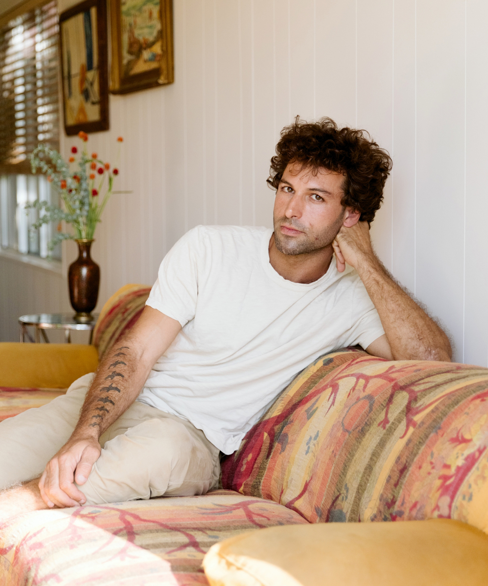 A person with curly hair wearing a white t-shirt and light pants sits on a patterned couch, resting their head on one hand and looking at the camera in this marc gabor-inspired scene. Framed paintings and a vase with flowers decorate the background.