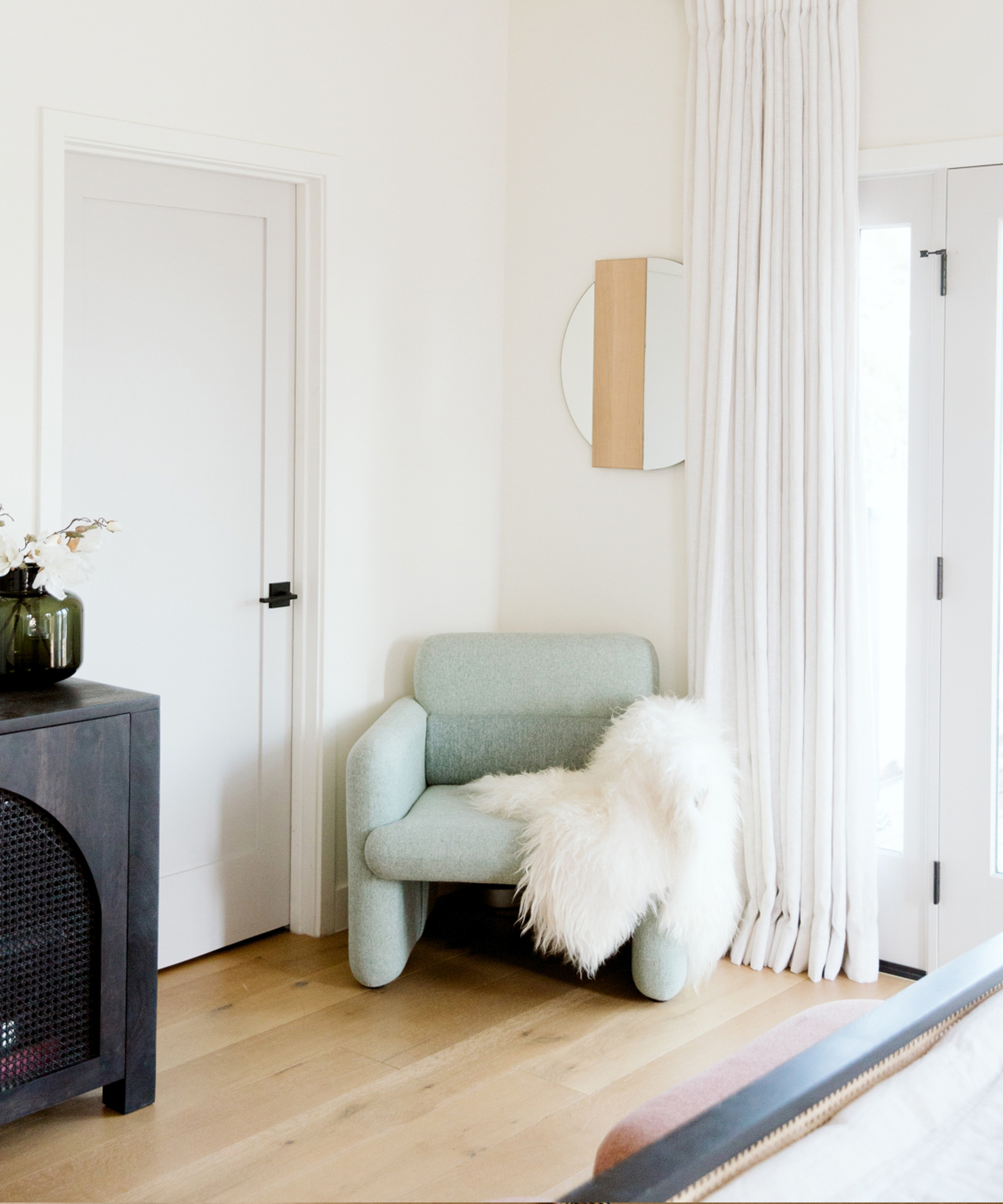 A minimalist room inspired by Jaclyn Johnson features a light green armchair draped with a white faux fur throw, light wood flooring, white walls, long white curtains, and a small black cabinet topped with a vase of flowers.