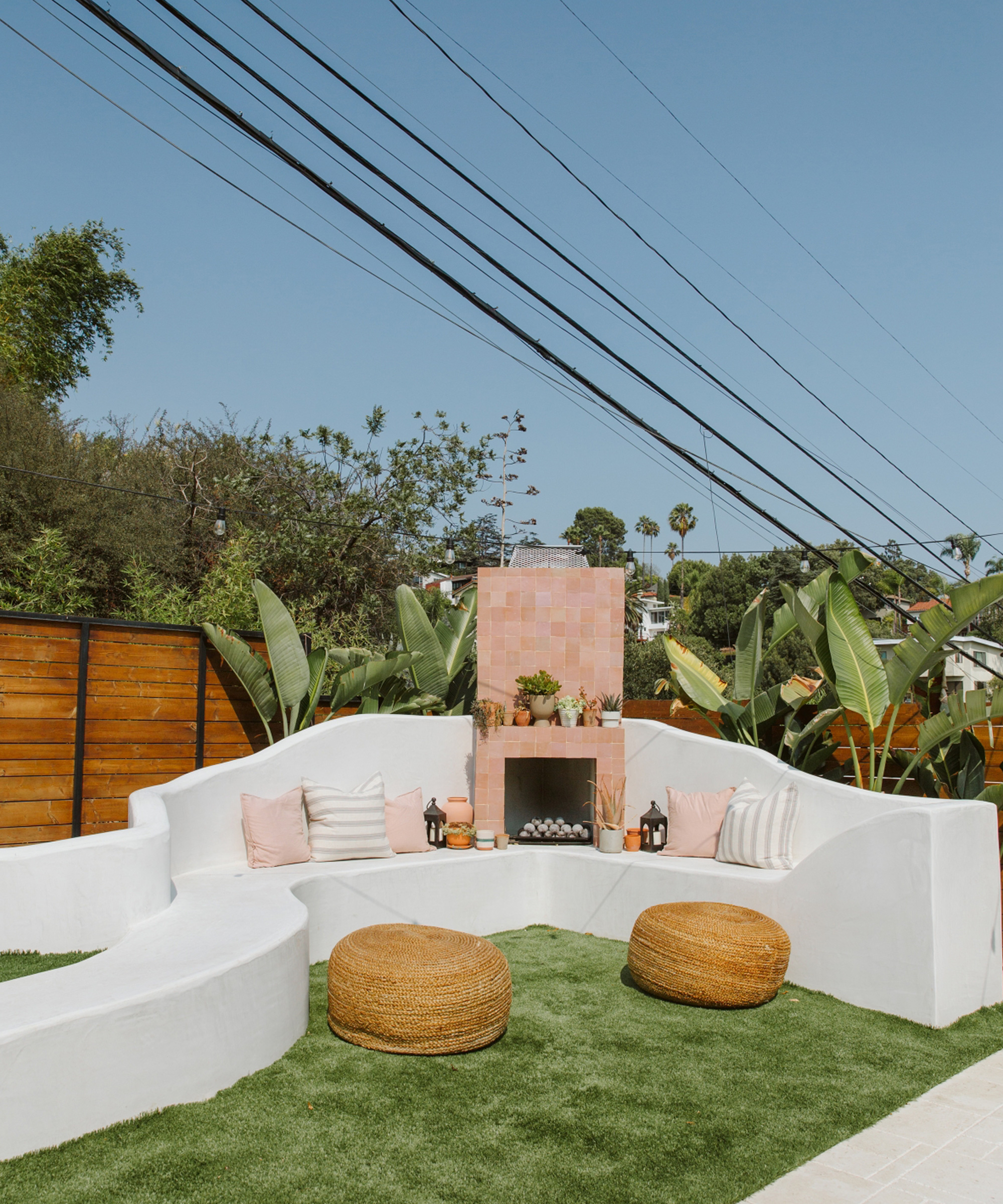 Outdoor patio styled by Jaclyn Johnson features curved white seating around a pink-tiled fireplace, plus pillows, lanterns, and lush potted plants. Woven ottomans rest on green artificial grass with a wooden fence as the backdrop.