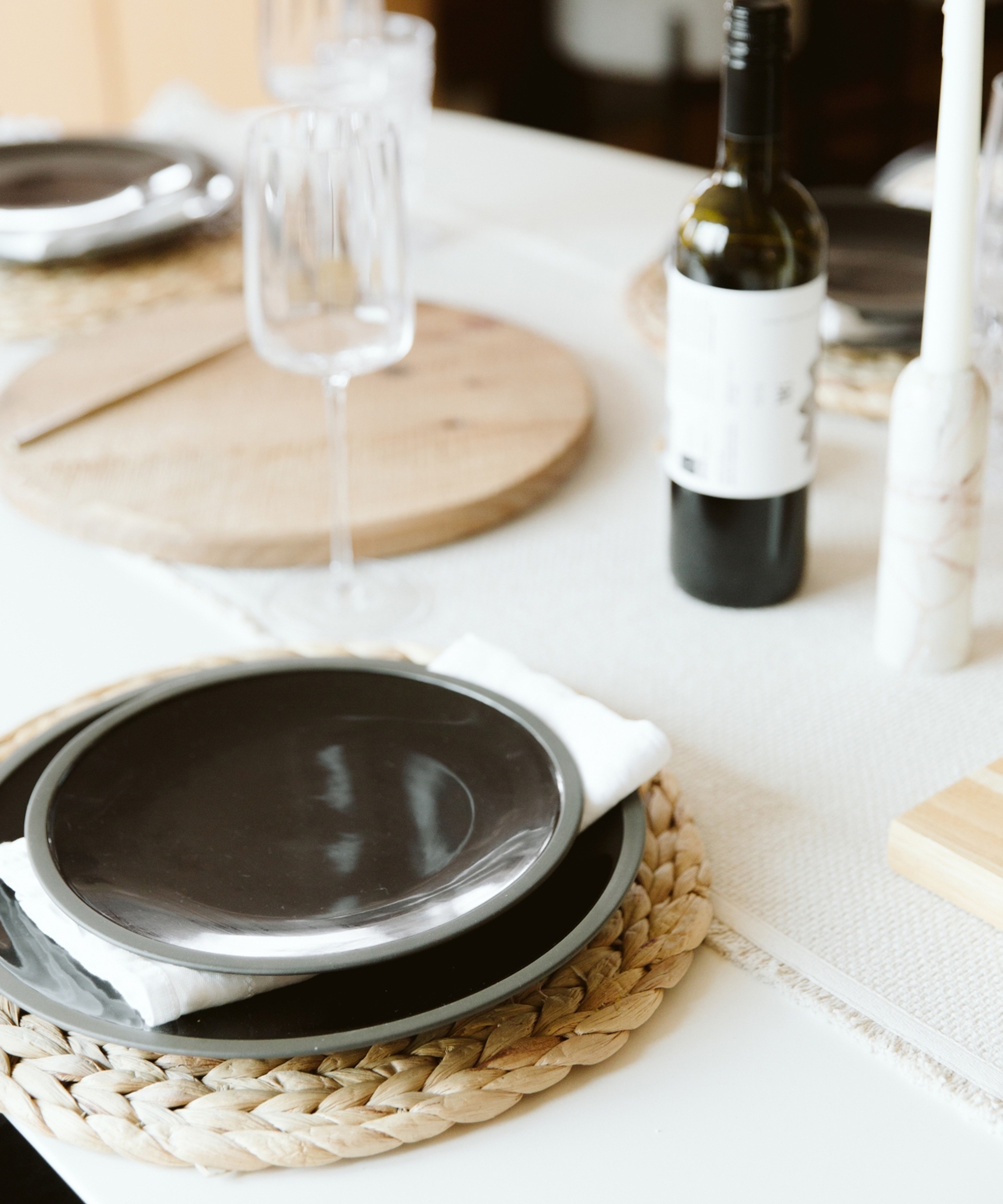 A close-up of a dining table set with black plates on woven placemats, white napkins, an empty wine glass, a bottle of wine, and round wooden boards on a light tablecloth styled in the elegant manner of Jaclyn Johnson.