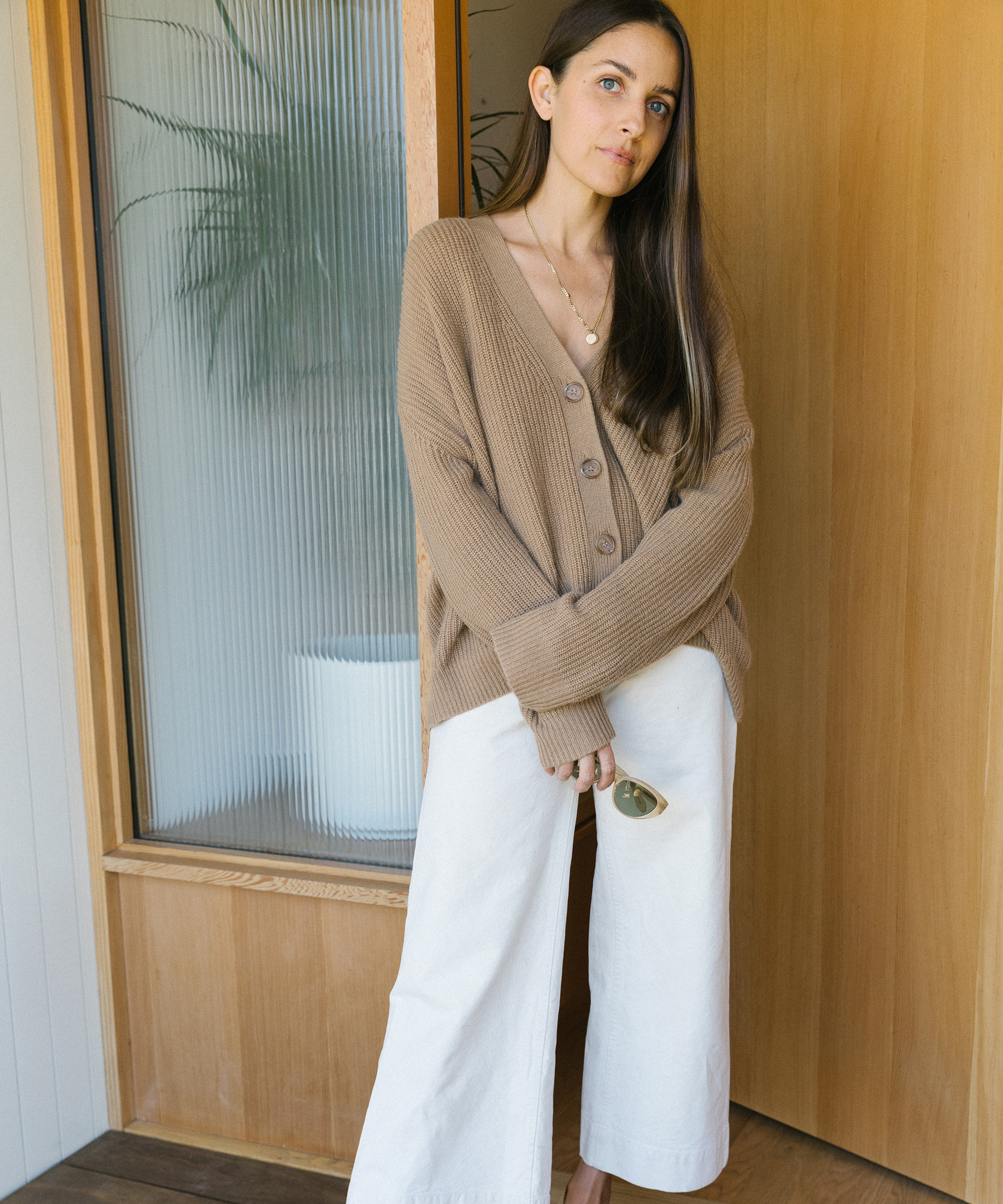 A woman stands indoors by a wooden door, wearing a cropped tan cardigan and wide-leg white pants. She holds sunglasses in one hand and looks calmly at the camera, with a green plant visible in the background.