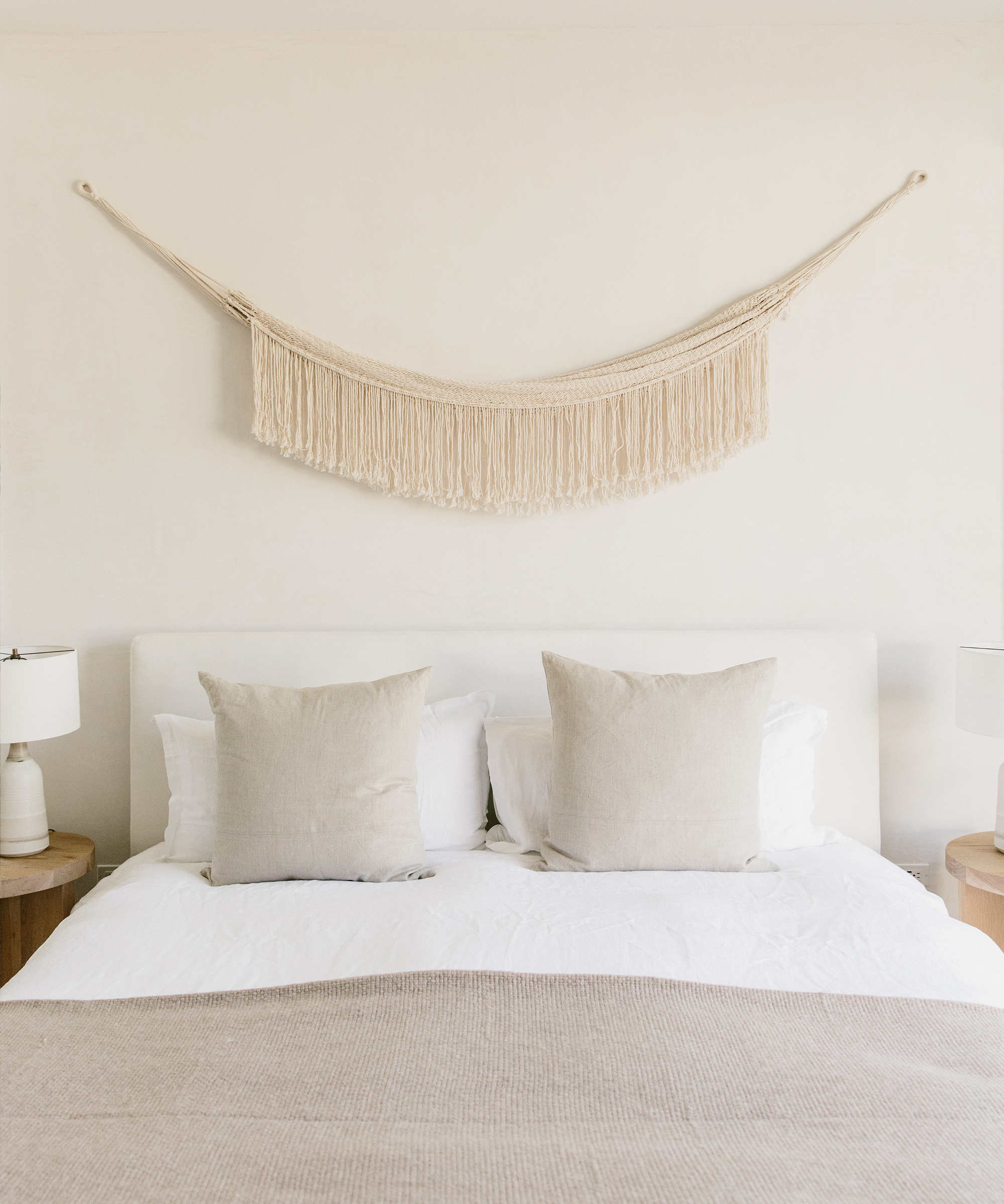 Bedroom with white linen duvet cover, oatmeal throws and linen pillows, and Jenni Kayne Woven Wall Hanging placed above the bed.
