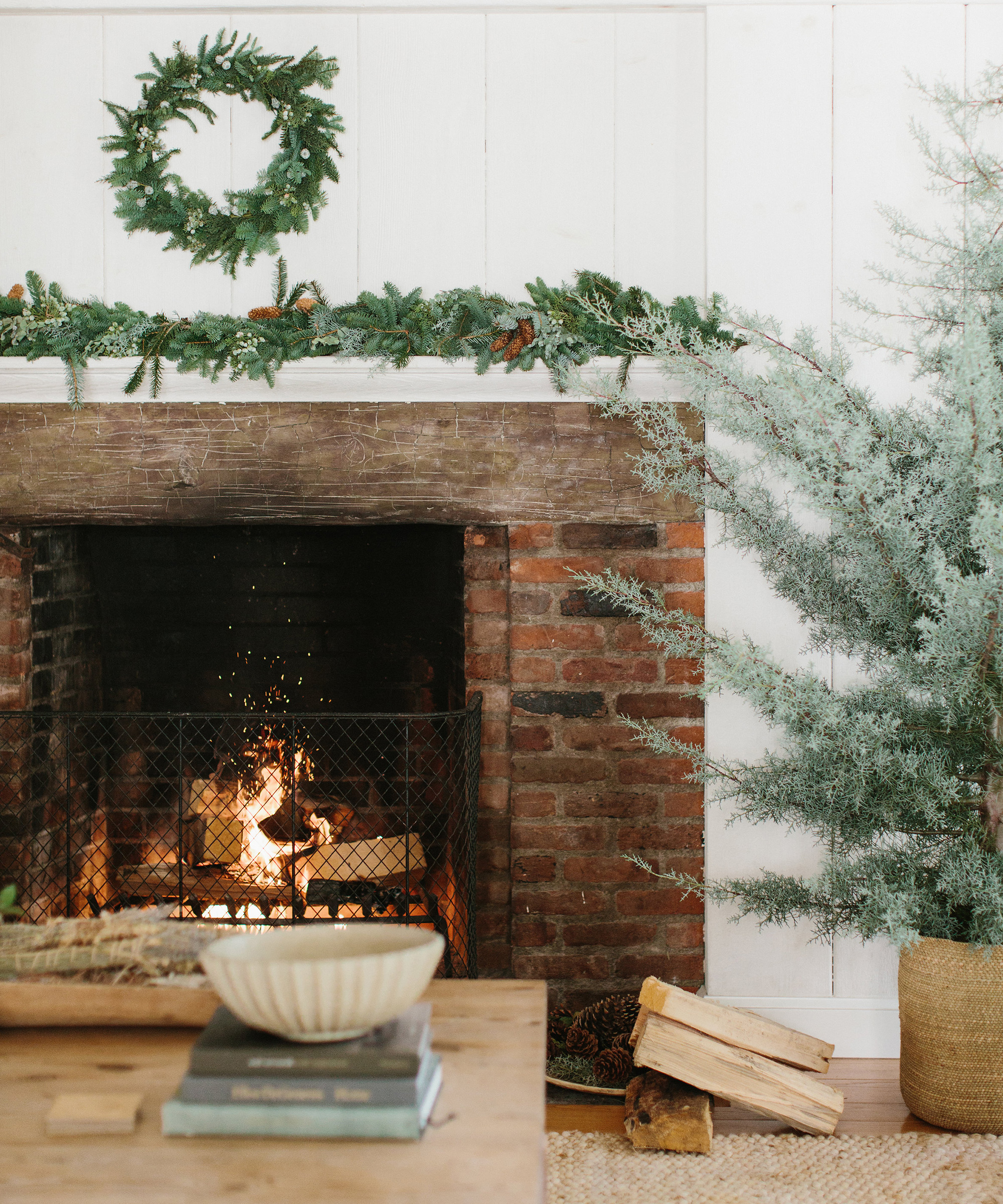 A cozy brick fireplace with a fire burning, decorated with a green garland and wreath. A stack of firewood, books, and holiday gift ideas sit on a wooden coffee table in a warmly lit living room beside a small evergreen tree.