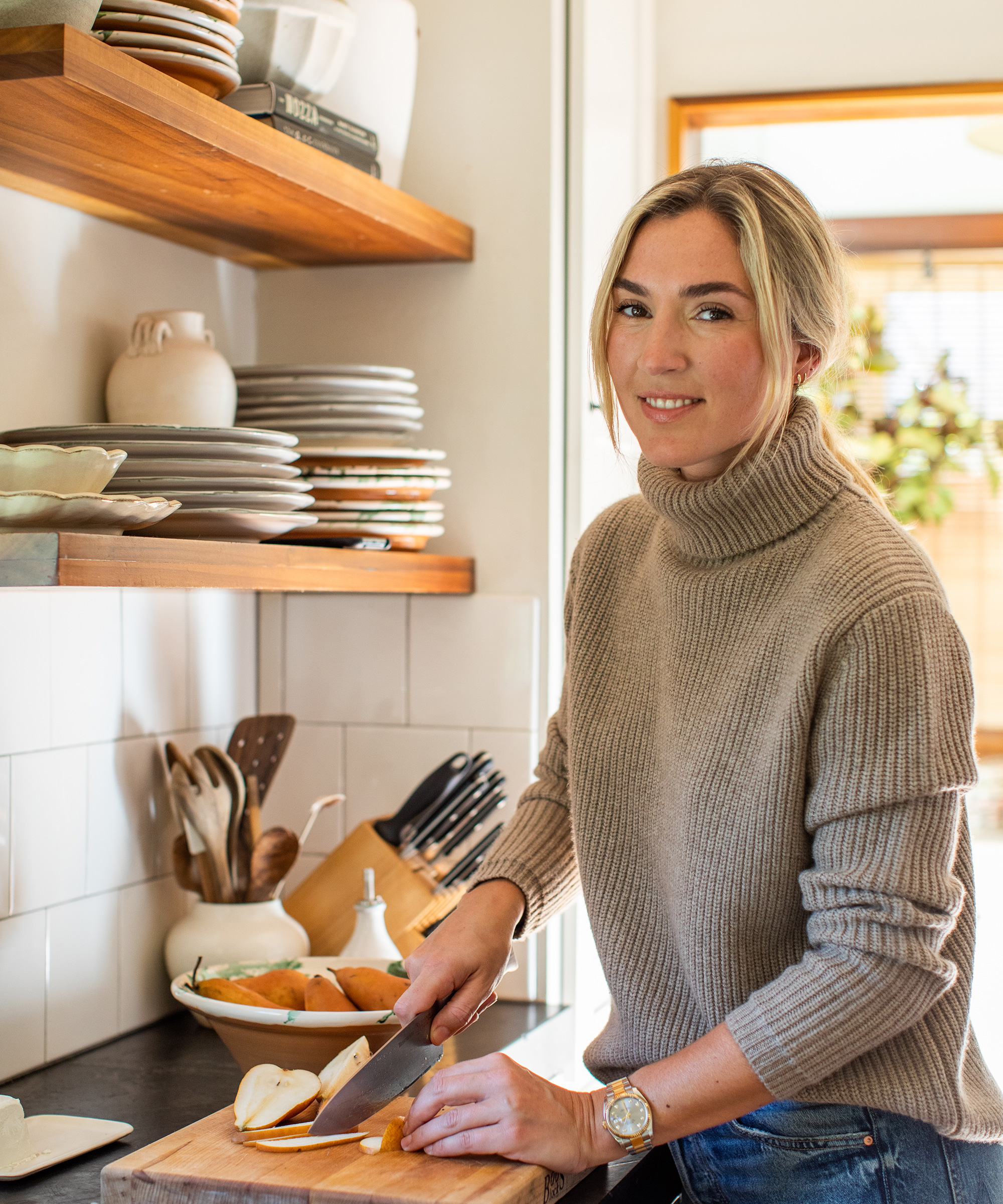 A woman in a beige turtleneck sweater smiles while slicing vegetables on a wooden cutting board, preparing one of the best fall recipes in a bright kitchen with shelves of plates and utensils in the background.