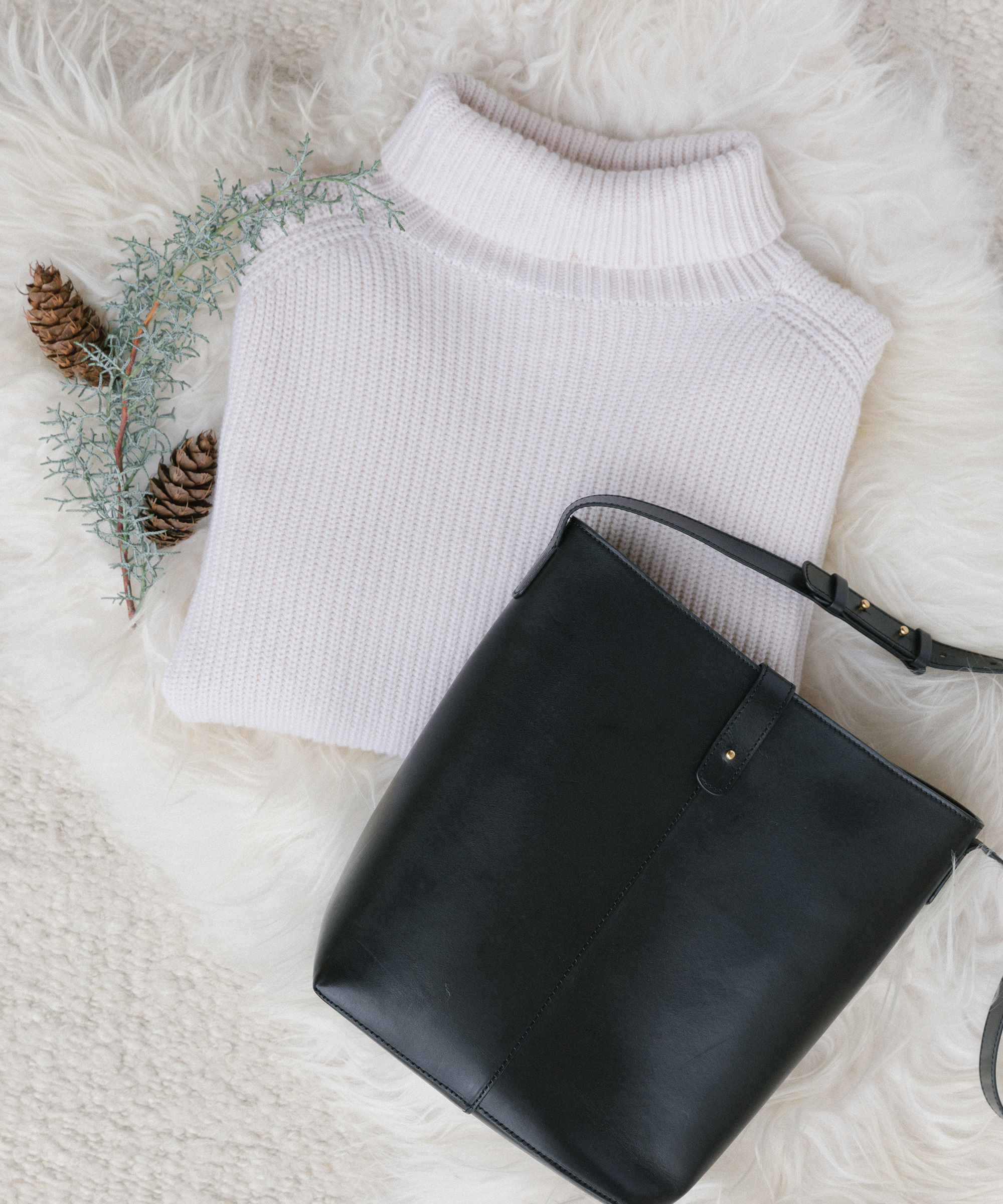 A folded white turtleneck sweater and a black leather handbag are placed on a white textured surface, next to a small sprig with pinecones and green foliage—perfect holiday gift ideas for any winter wardrobe.