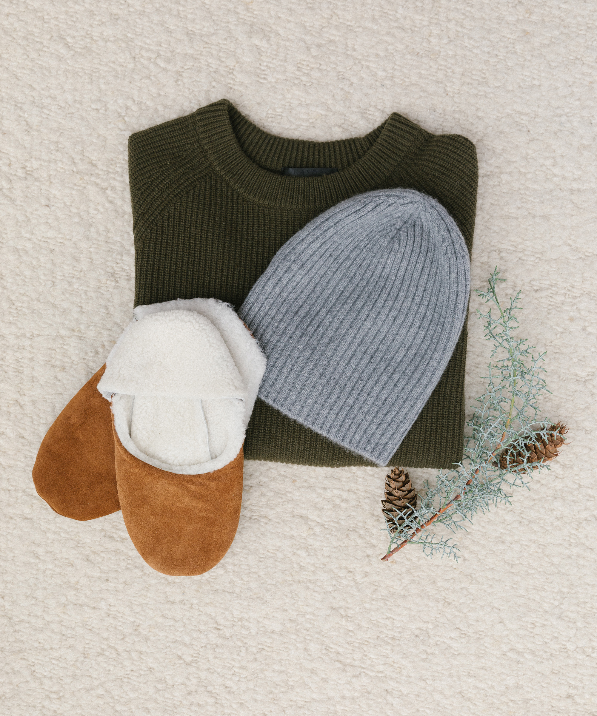 A folded green sweater, a gray knit beanie, brown slippers with white lining, and a small pine branch with pinecones arranged on a cream-colored textured surface—perfect for cozy holiday gift ideas.