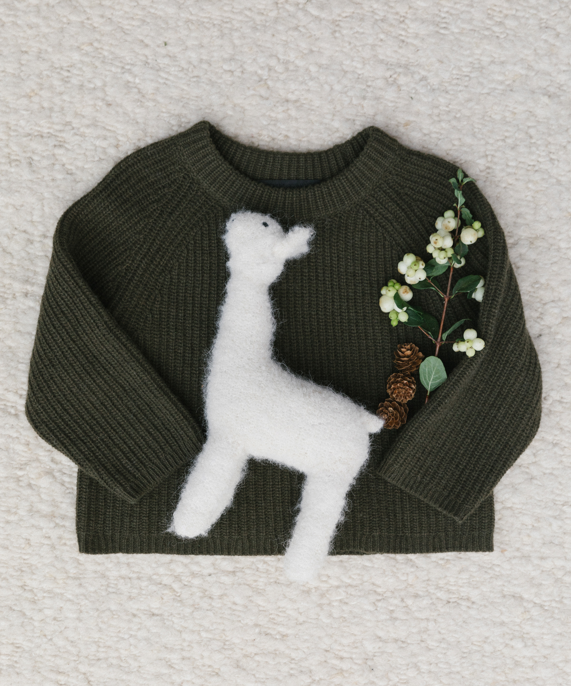 A dark green knitted sweater featuring a large, fuzzy white animal design—possibly a llama or alpaca—on the front. A small branch with white berries and pinecones rests on it, making it perfect for cozy holiday gift ideas.