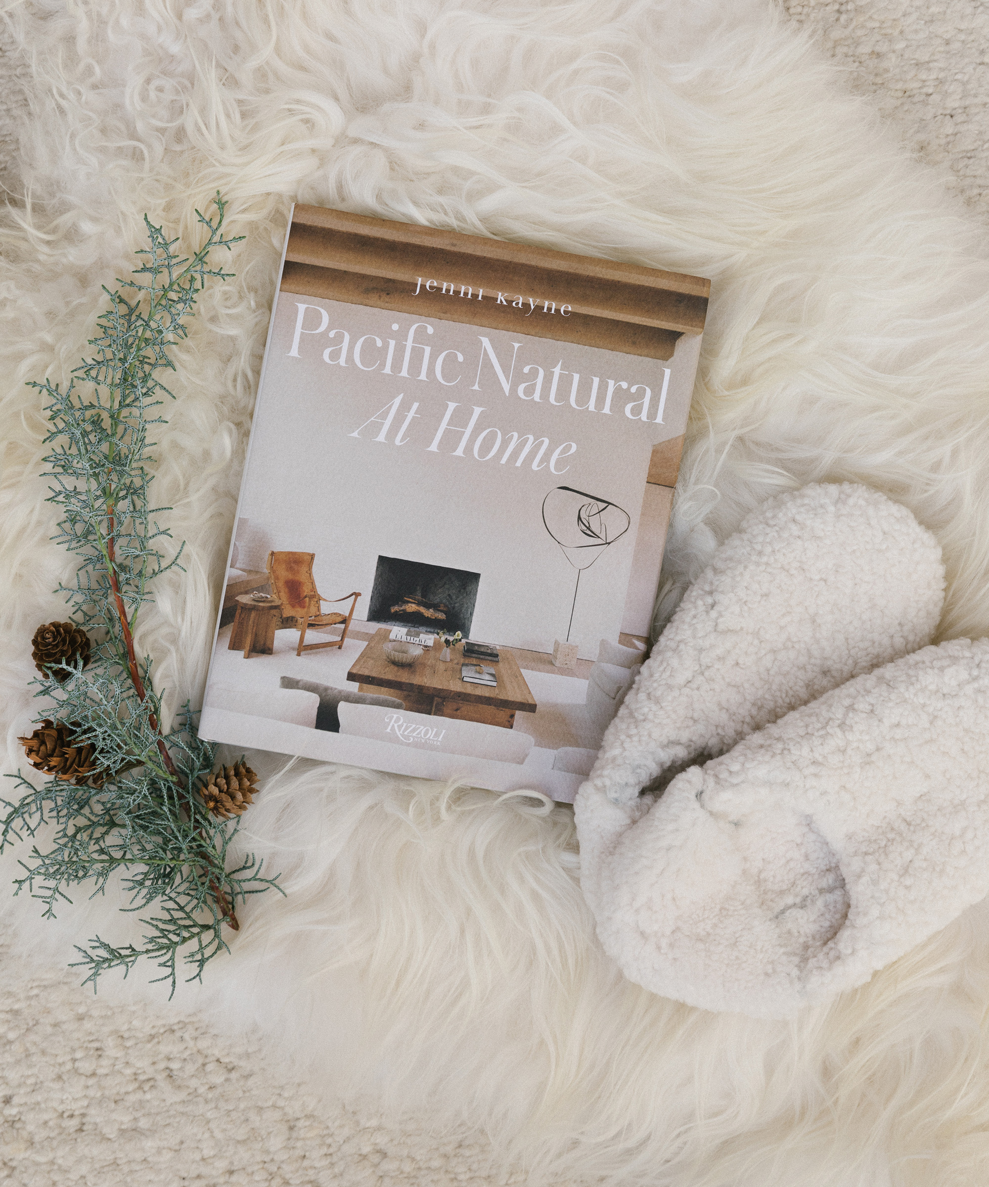 A book titled Pacific Natural At Home rests on a white fluffy surface beside a sprig of greenery with pinecones and a pair of fuzzy white slippers, offering cozy holiday gift ideas for seasonal relaxation.