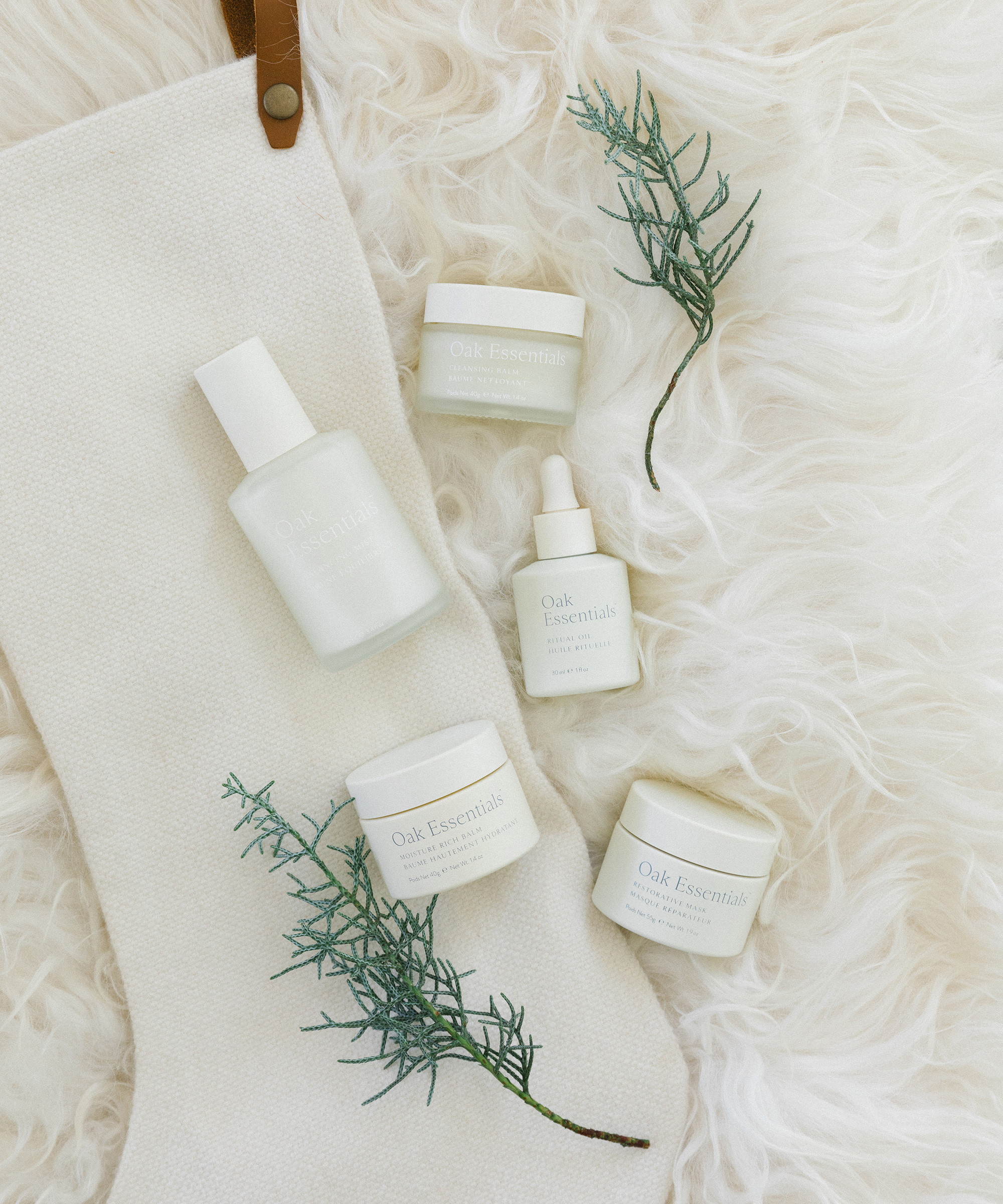 A cream-colored stocking and five Oak Essentials skincare products are arranged on a fluffy white surface with two sprigs of greenery, making it perfect for holiday gift ideas.