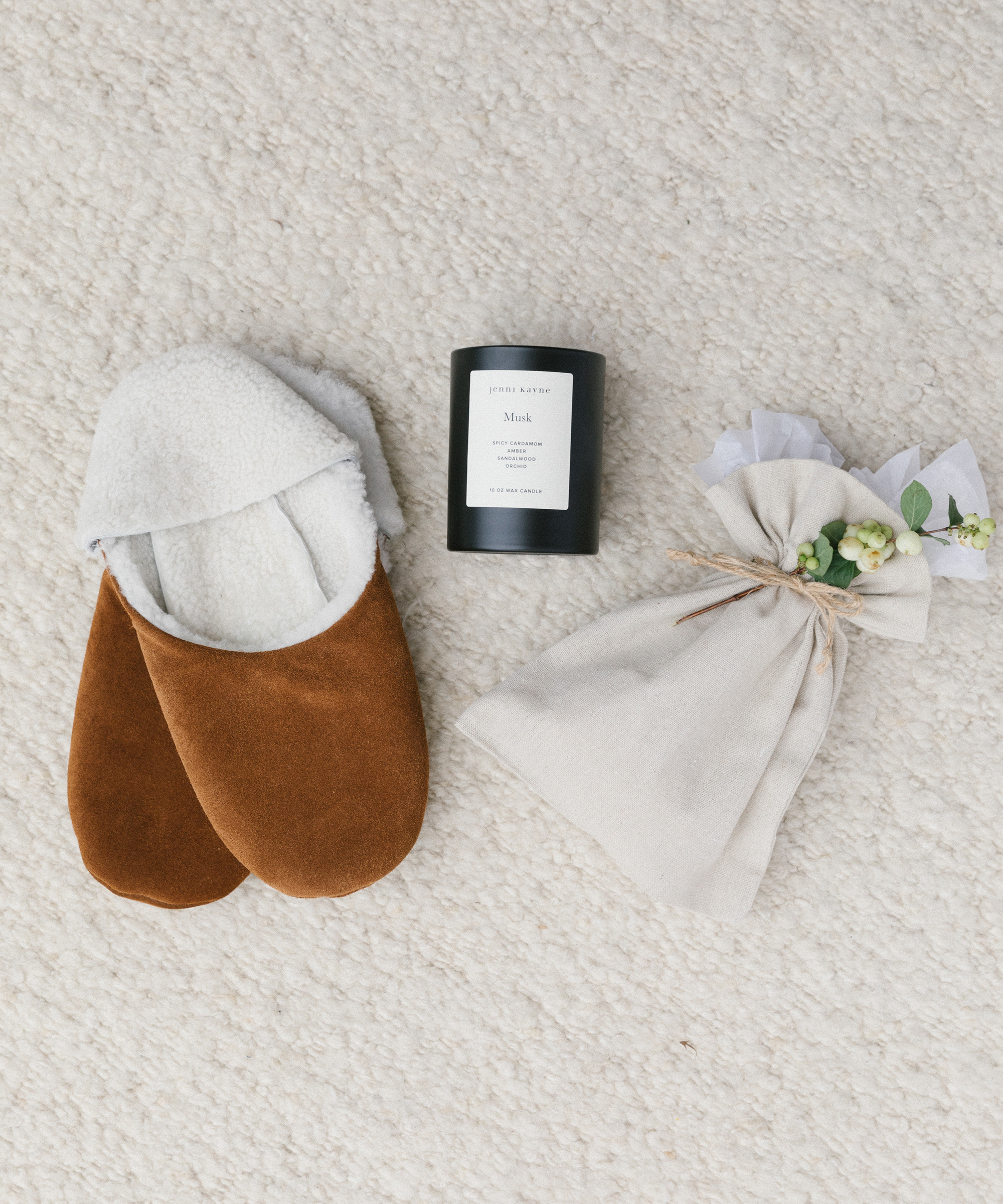 A pair of brown slippers, a black candle labeled Musk, and a fabric pouch tied with twine and green berries—thoughtful touches arranged on a light textured carpet, perfect for unique holiday gift ideas.
