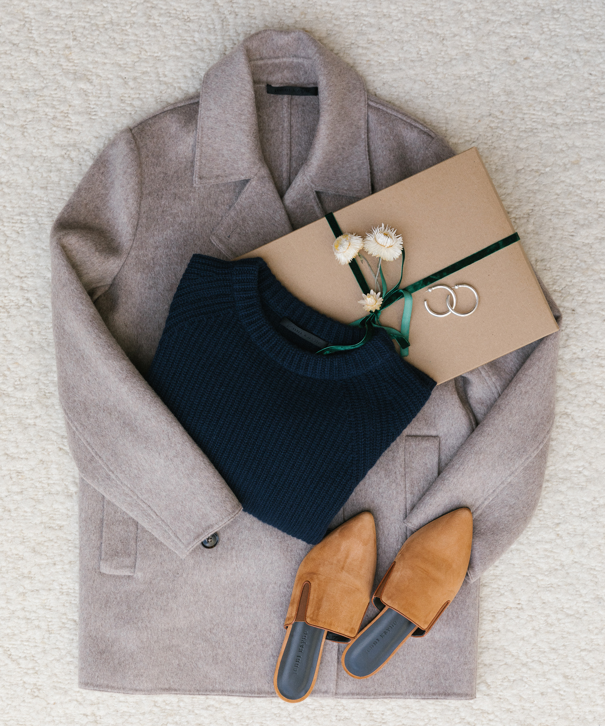 A beige wool coat, a navy blue sweater, tan suede mules, and a gift wrapped in brown paper with flowers and green ribbon are arranged on a light textured surface—perfect inspiration for holiday gift ideas.