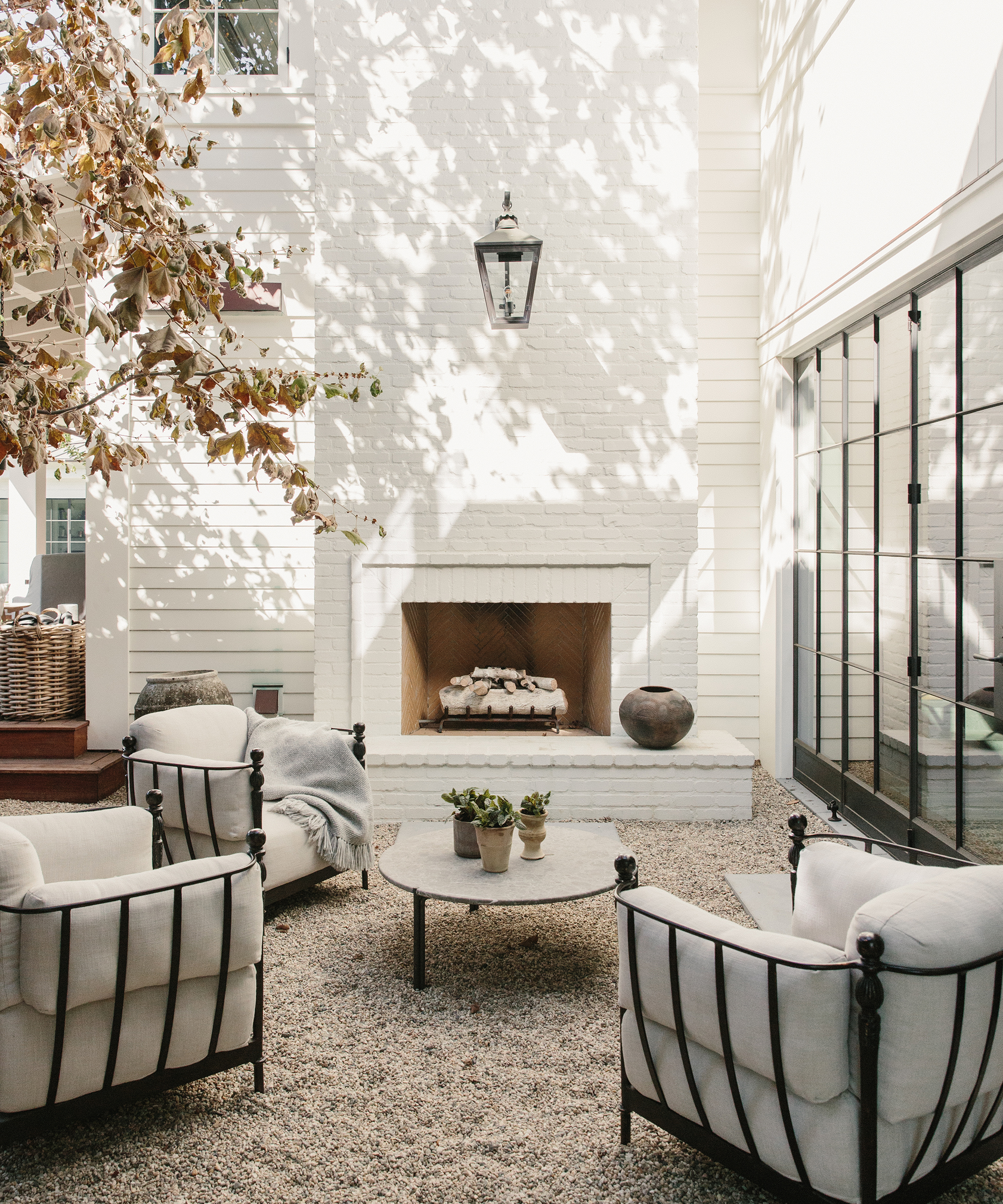 A cozy outdoor patio by m. elle design features three cushioned armchairs around a round coffee table, a white brick fireplace, potted plants, and a tree casting dappled shadows on the white walls.