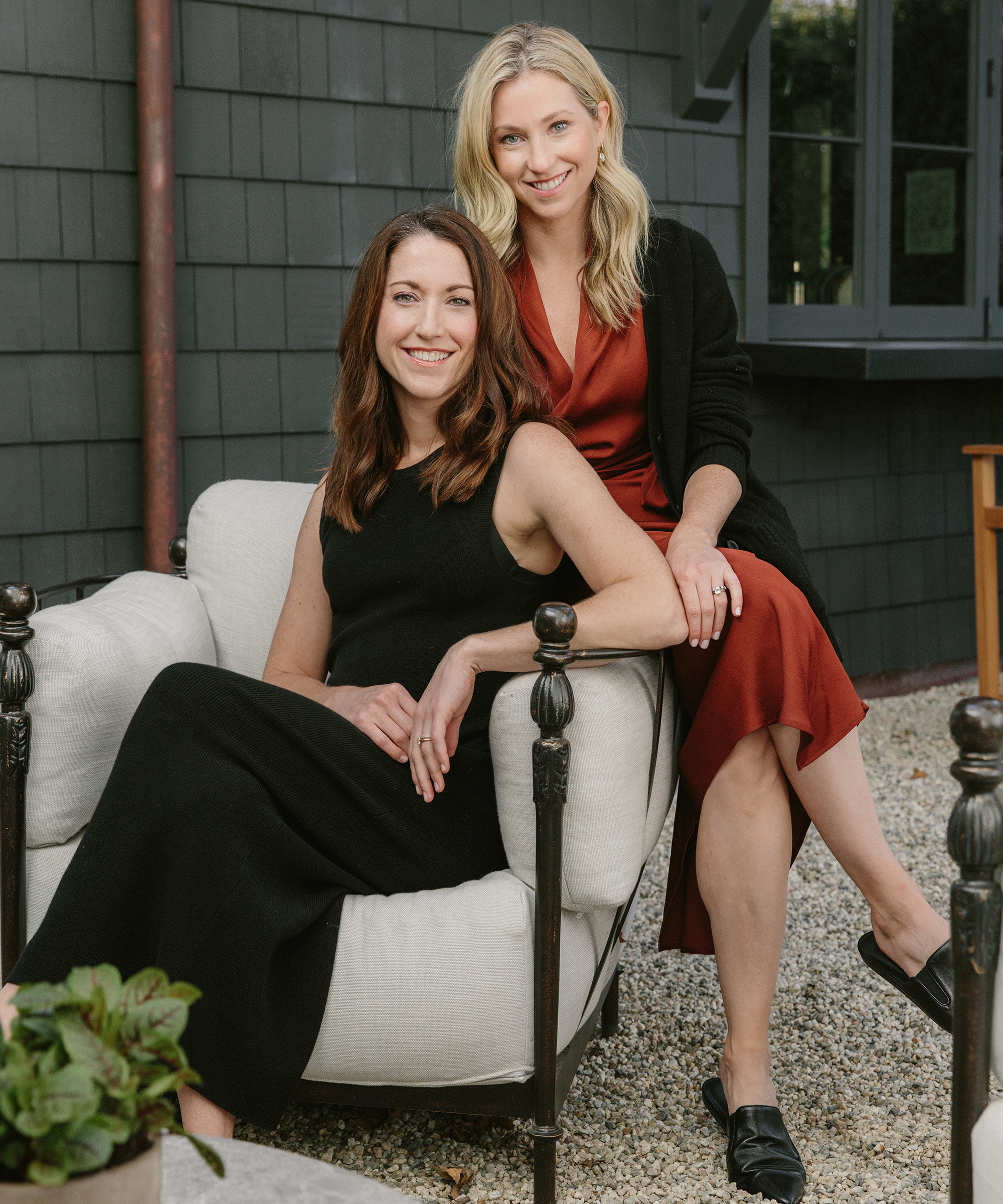 Two women pose outdoors in elegant m. elle design attire. One sits on a cushioned chair in a sleeveless black dress, smiling, while the other stands beside her in a rust-colored dress and black cardigan, hand resting supportively on her shoulder.