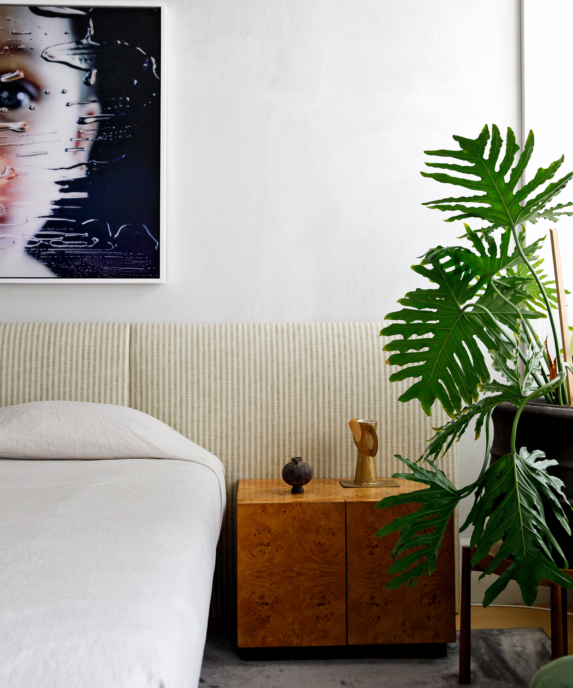 A modern bedroom with a light-colored bed, a wooden nightstand holding decor items, a large leafy plant, and a framed abstract face artwork on a white wall inspired by Jane Keltner de Valle’s signature style.