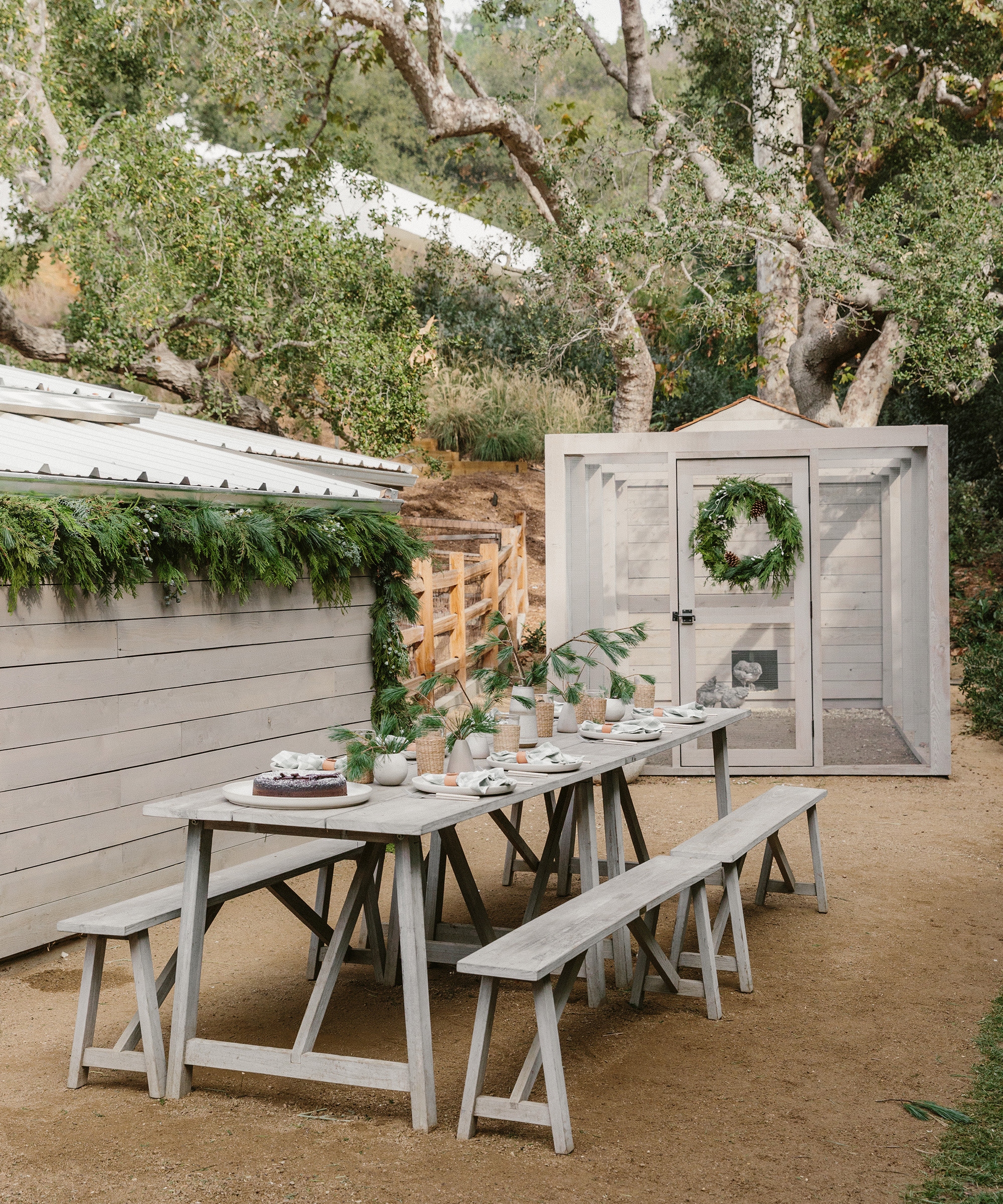A rustic outdoor dining area with a long wooden table set for a meal, featuring holiday table decor. Greenery adorns the shed and a wreath hangs on a small structure, all beneath leafy trees and surrounded by benches.