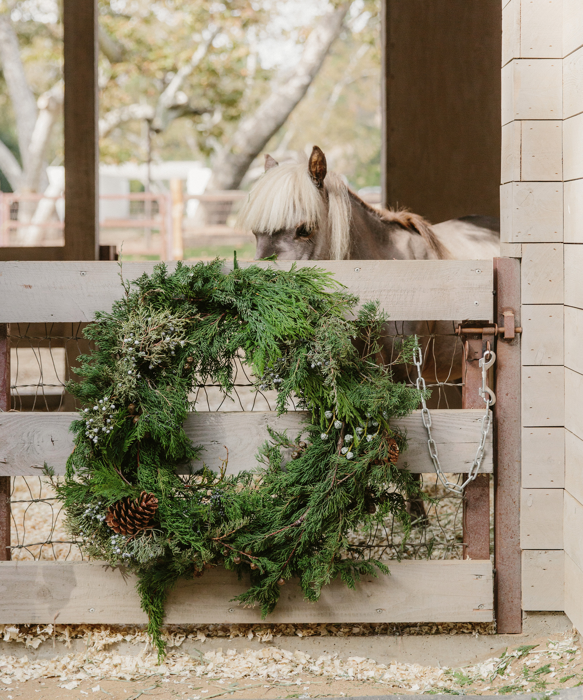 A small pony stands behind a wooden gate decorated with a large green holiday wreath adorned with pinecones and foliage, echoing the charm of holiday table decor in a rustic barn setting.