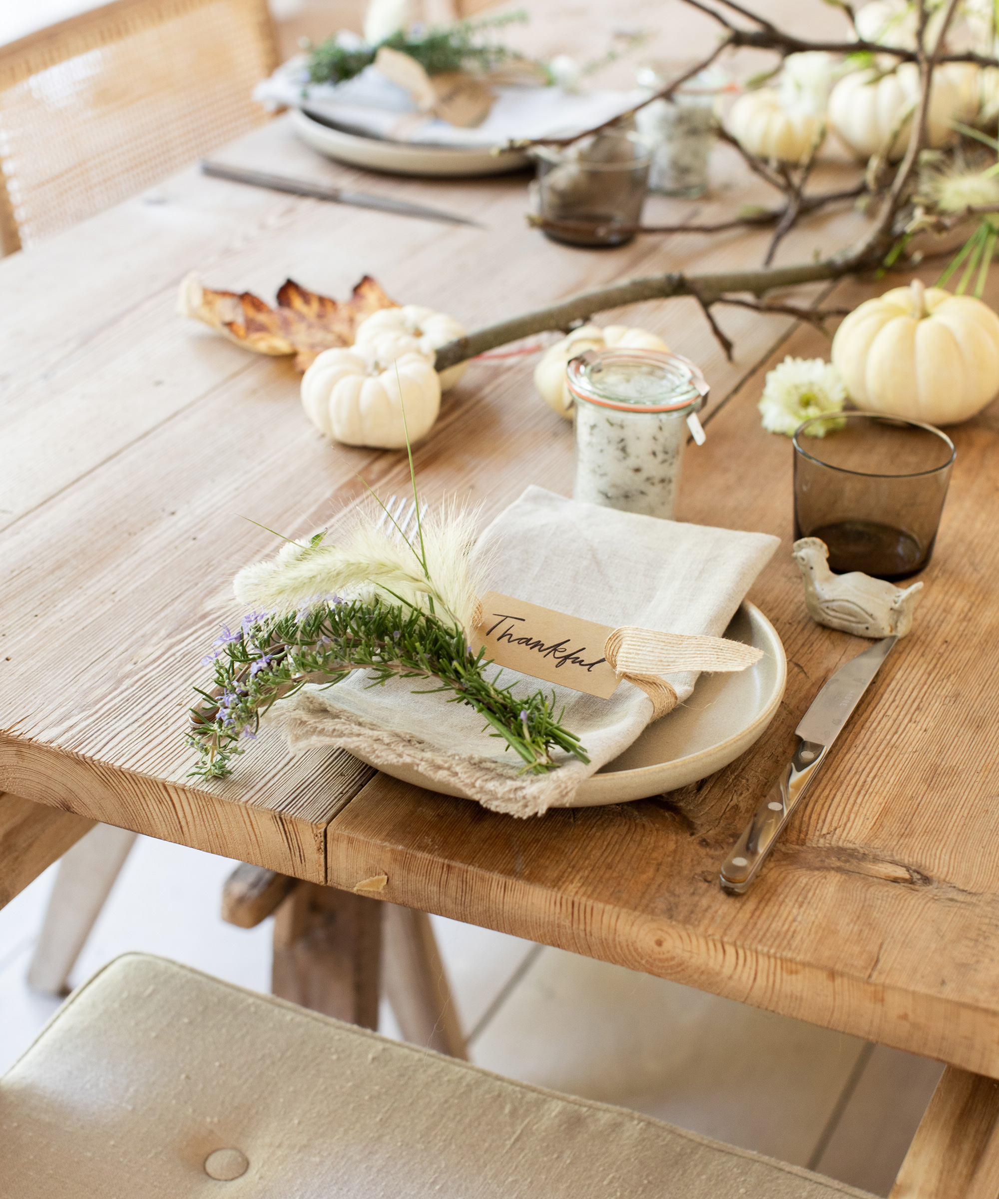 A rustic wooden table set for a meal with a plate, linen napkin, silverware, a glass, a jar, white pumpkins, branches, and a Thankful card is beautifully decorated with greenery and flowers—perfect inspiration for your Thanksgiving recipe ideas.