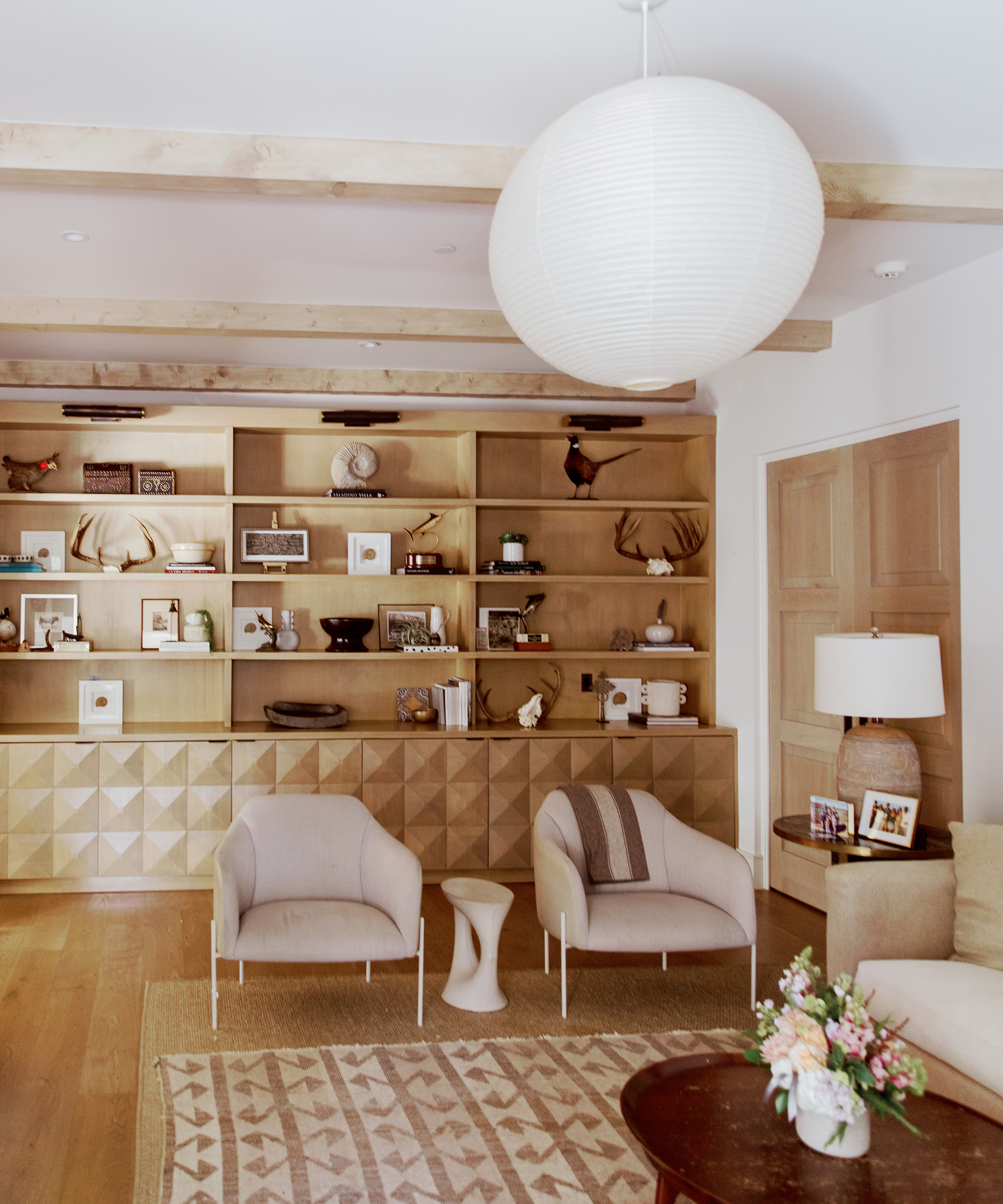 A modern living room with light wood floors designed by Emily Seiders features geometric wooden shelves displaying decor, two beige armchairs, a side table, a patterned rug, and a round paper pendant light overhead. A small flower arrangement sits on the coffee table.
