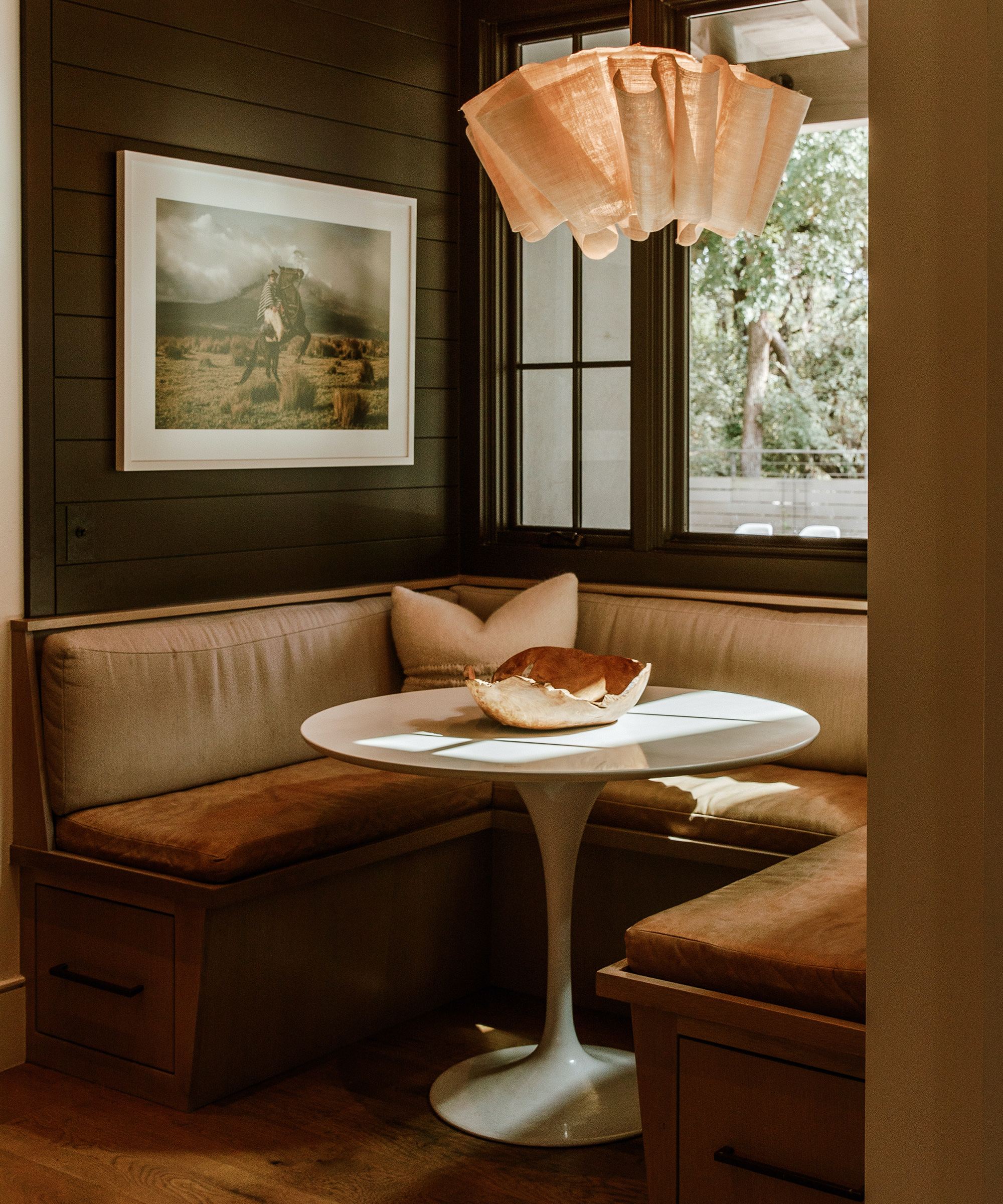 A cozy breakfast nook designed by Emily Seiders features brown cushioned benches, a round white table, a decorative bowl, a beige pillow, a Western-themed photograph on the wall, and a sculptural fabric pendant light by the window.