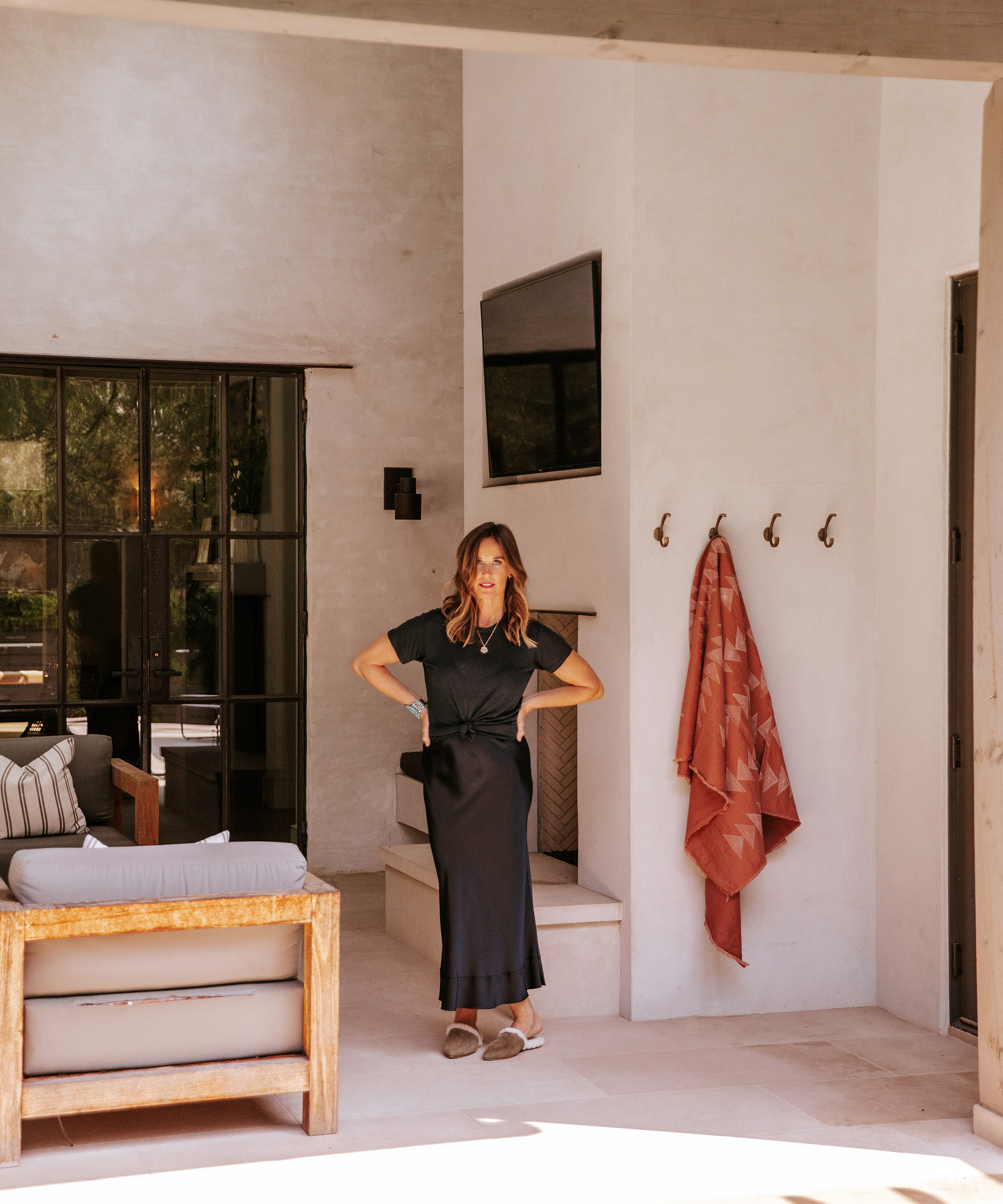 Emily Seiders stands with hands on hips in a sunlit, modern patio area. She wears a black dress and sandals. Behind her are a mounted TV, hooks with a towel, a couch, large window, and door.