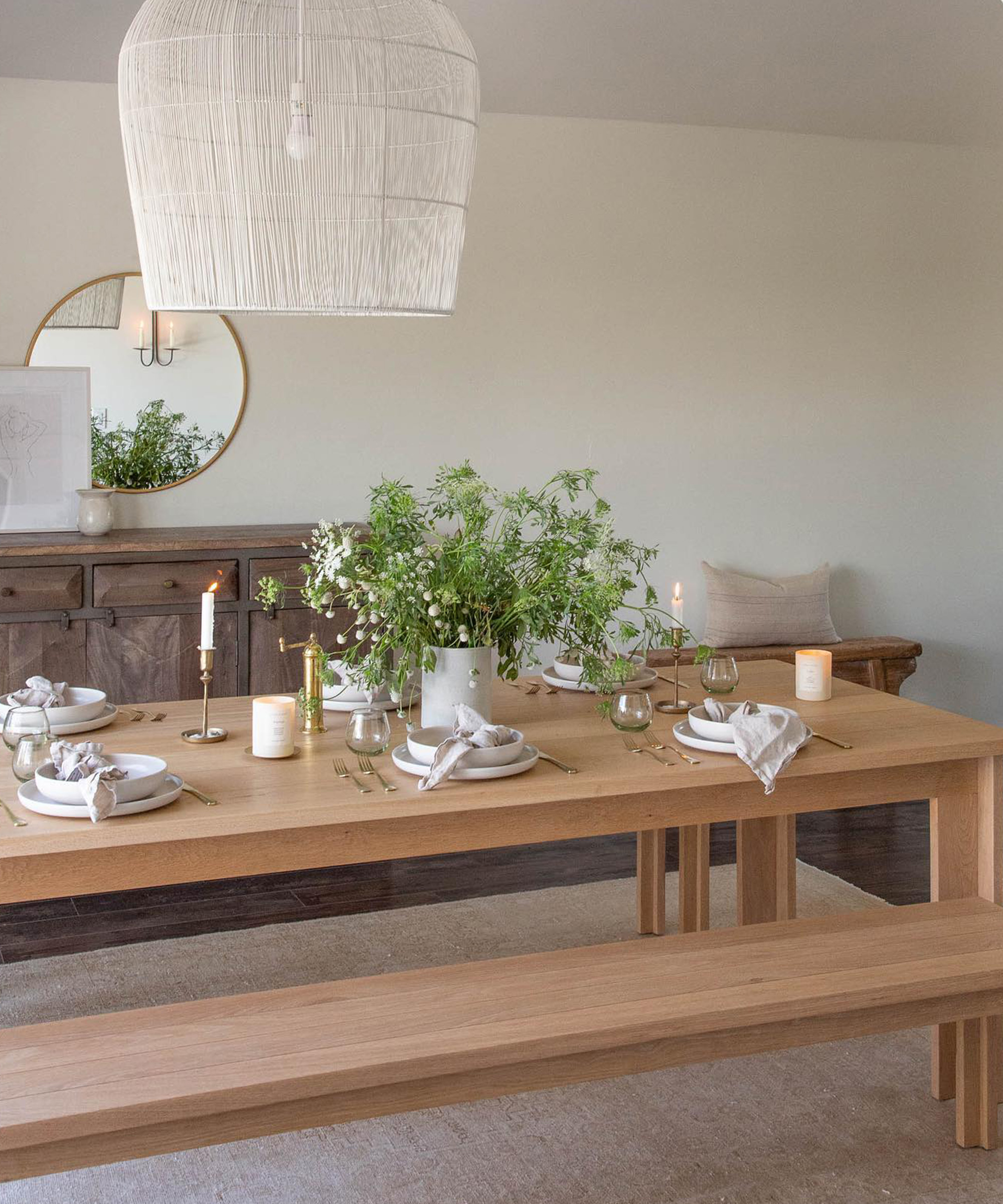 A light wood dining table with benches is set with white dishes, candles, and a large bouquet of greenery. A white woven pendant light hangs above, offering bright, airy interior design inspiration alongside a sideboard with a round mirror.