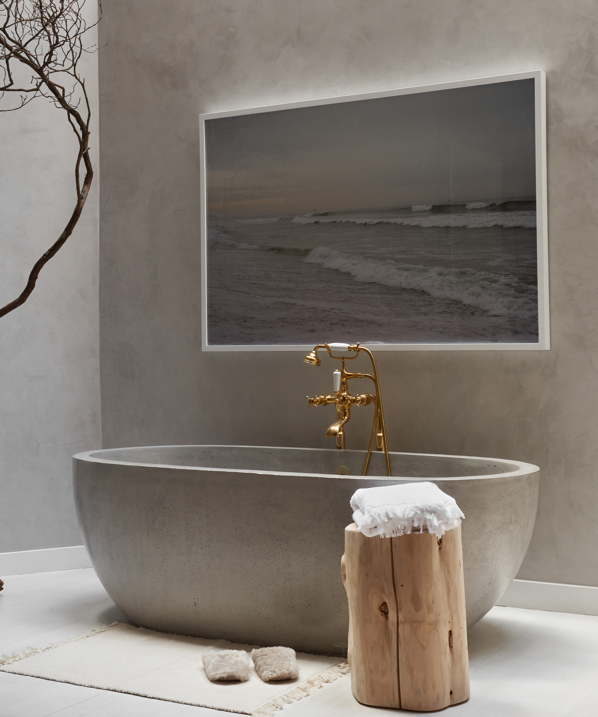 A modern bathroom with a gray stone bathtub, gold fixtures, and a wooden stool topped with a white towel—all styled like an NYC furniture store—features beige slippers on a rug and a large framed ocean photograph on the wall.