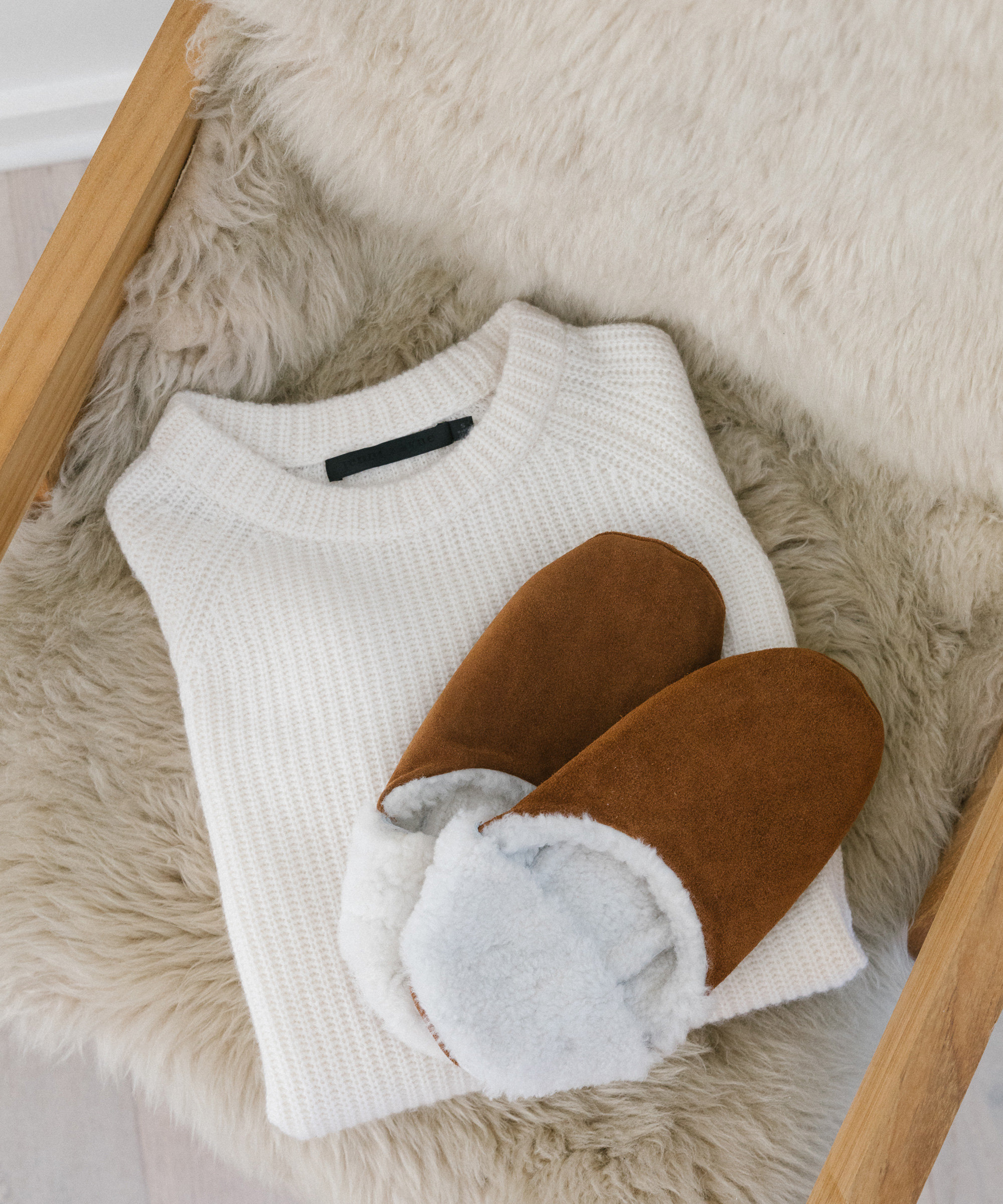 A folded white knit sweater and a pair of brown, fuzzy-lined slippers rest on a beige faux fur-covered chair—cozy essentials that make perfect valentines day gifts.