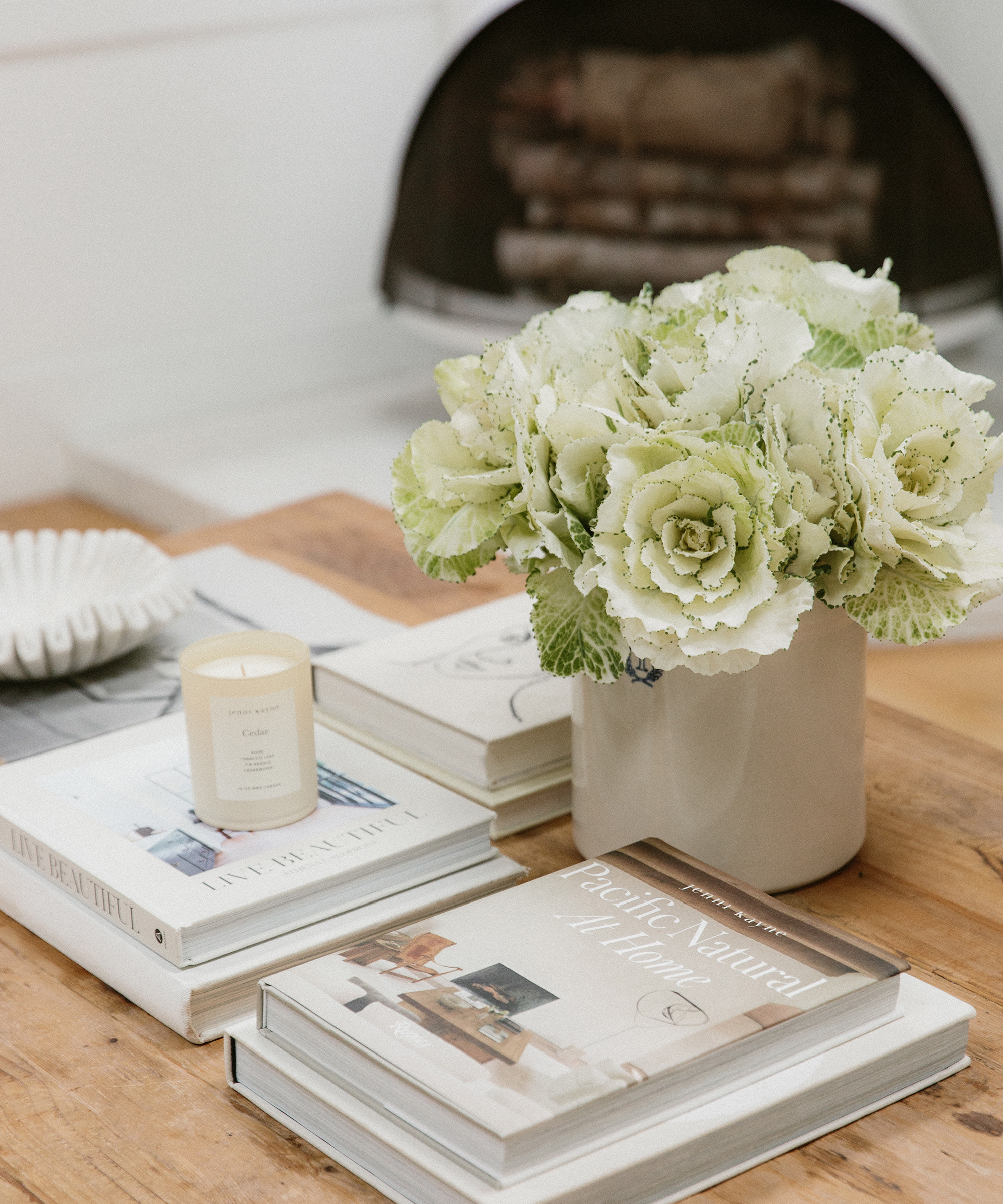 A cozy tabletop scene styled by Cydney Edwards features a vase of green and white ornamental cabbage flowers, a lit candle, and neatly stacked books, including Pacific Natural at Home, on a wooden coffee table.