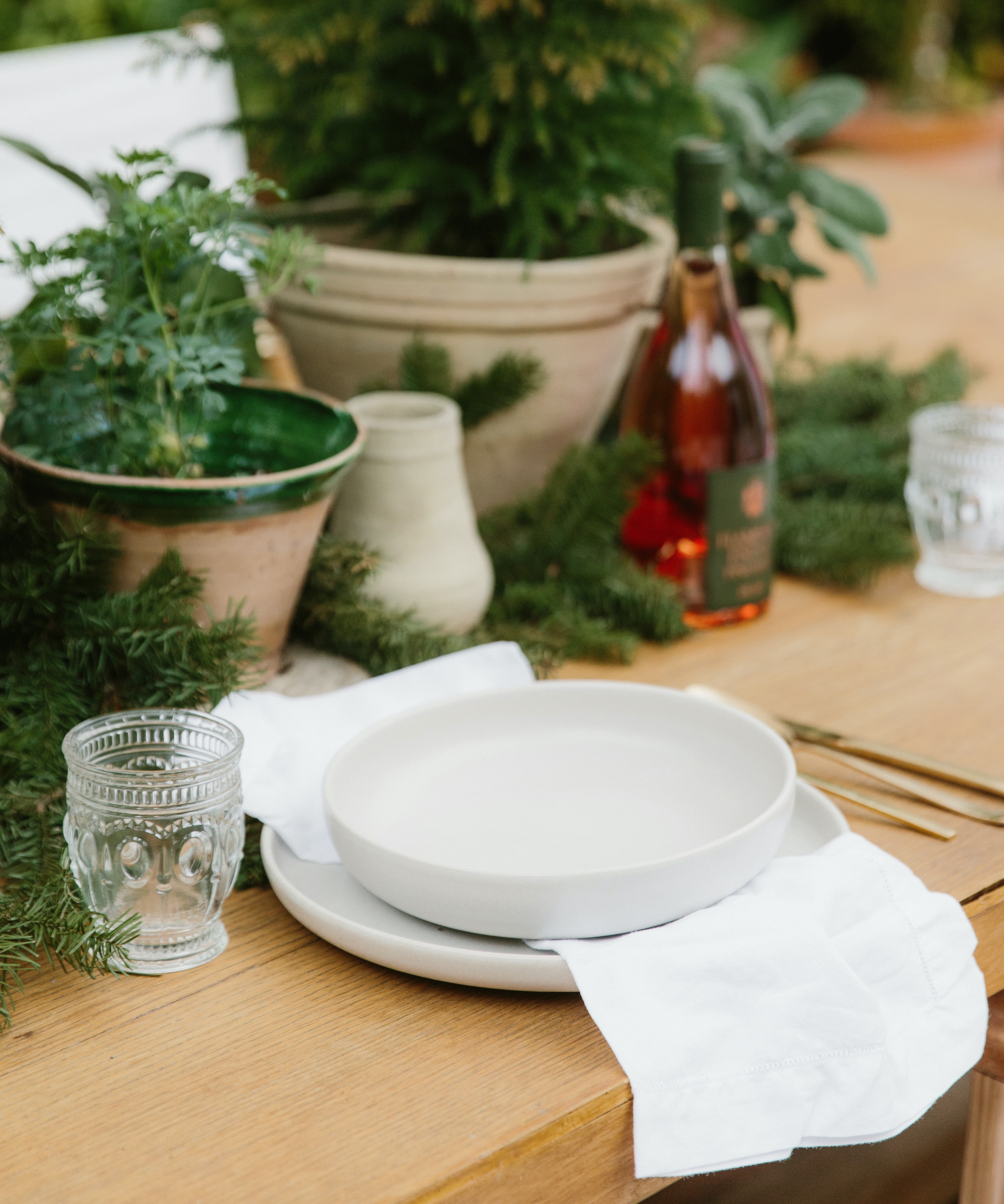 A wooden table set with white plates, a white napkin, a clear glass, and surrounded by green plants and a bottle of rosé wine. Cydney Edwards creates an inviting outdoor atmosphere that feels both fresh and welcoming.