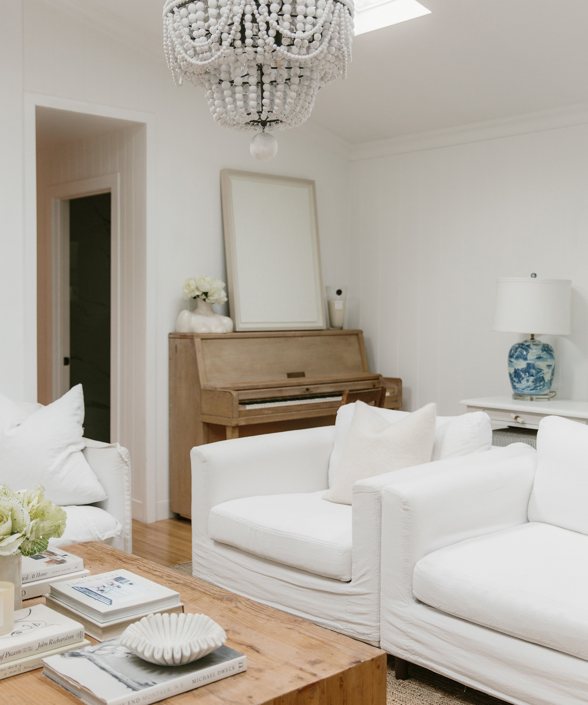 A bright, airy living room by Cydney Edwards features white armchairs, a wooden coffee table with books, a light wood piano with a mirror above it, and a blue-and-white lamp on a side table. A beaded chandelier hangs from the ceiling.