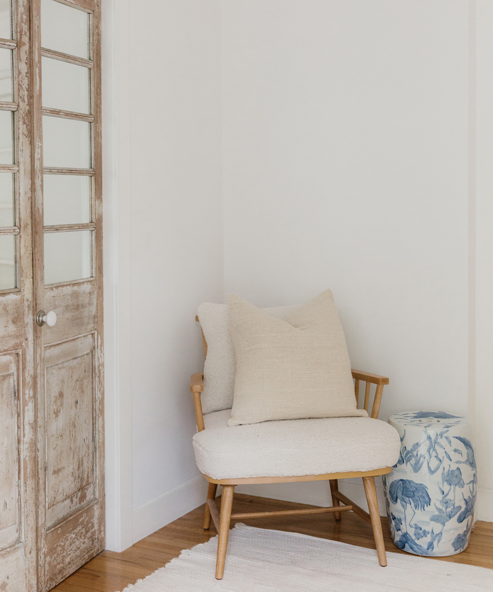 A cozy wooden chair with beige cushions sits in a corner next to a blue and white ceramic vase, styled by Cydney Edwards, against white walls and beside a rustic wooden door with glass panels.