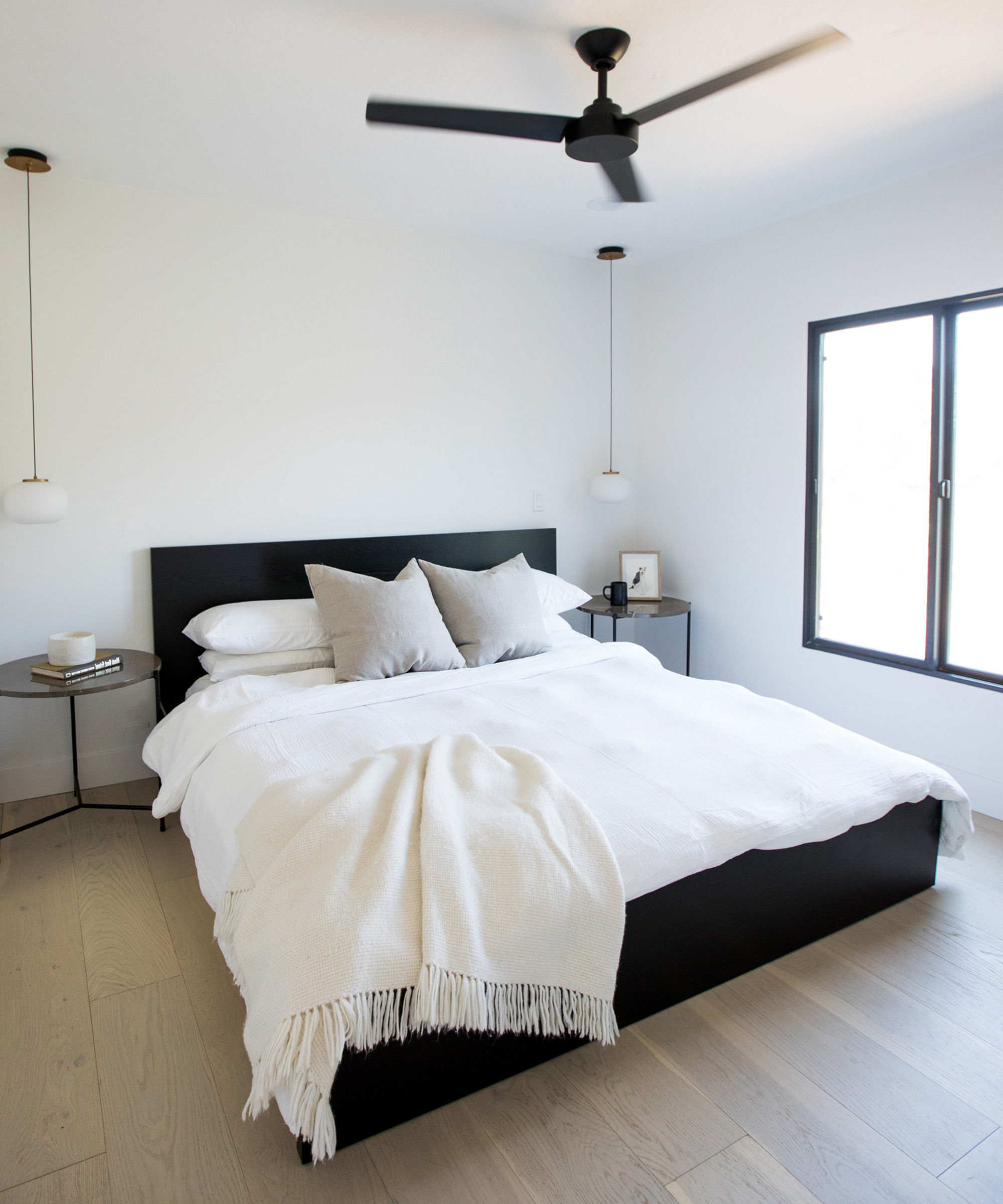 Minimalist bedroom by lou home featuring a black bed frame, white bedding, two gray pillows, round pendant lights, nightstands on each side, a window with natural light, and a black ceiling fan above.